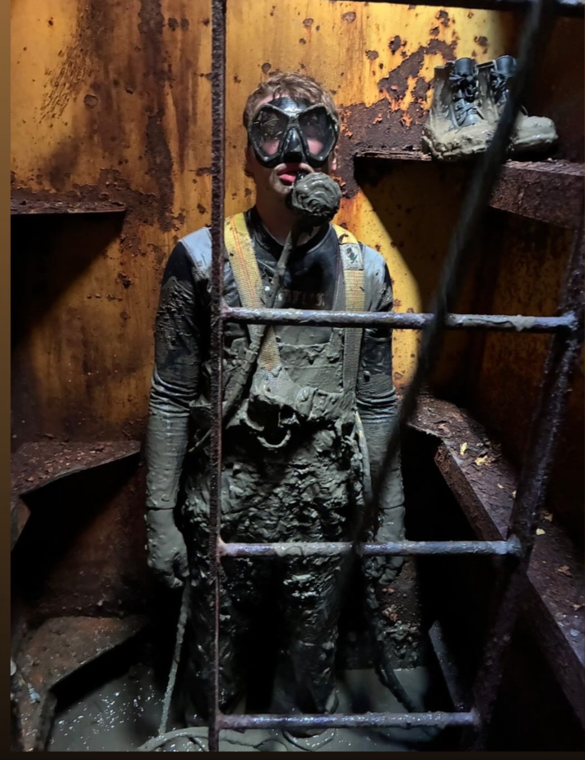 Person covered in mud wearing a mask and safety gear inside a rusty, confined space.