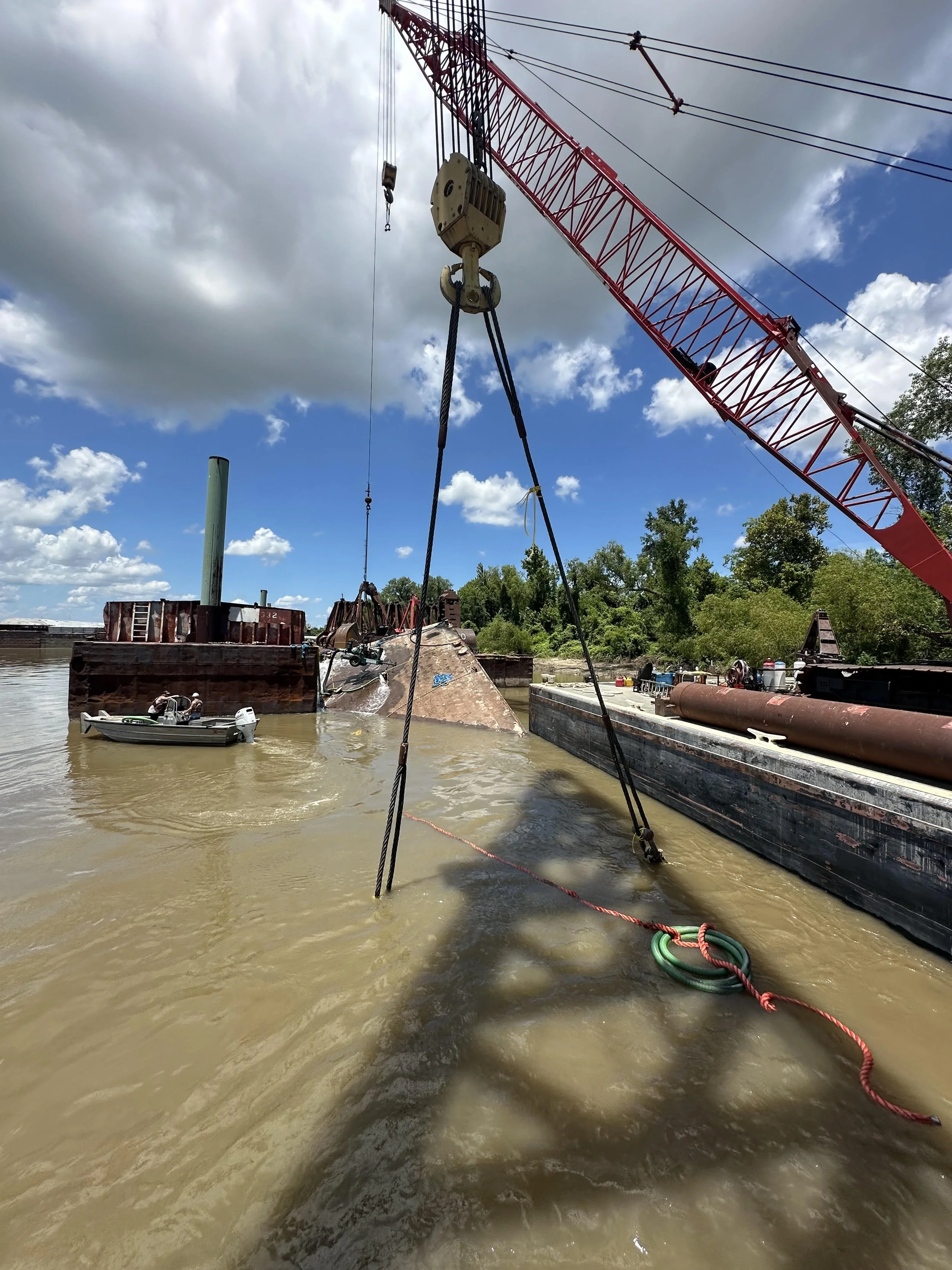 A large crane lifting a rusted shipwreck in a river, with a small boat nearby and trees in the background, under a partly cloudy sky.