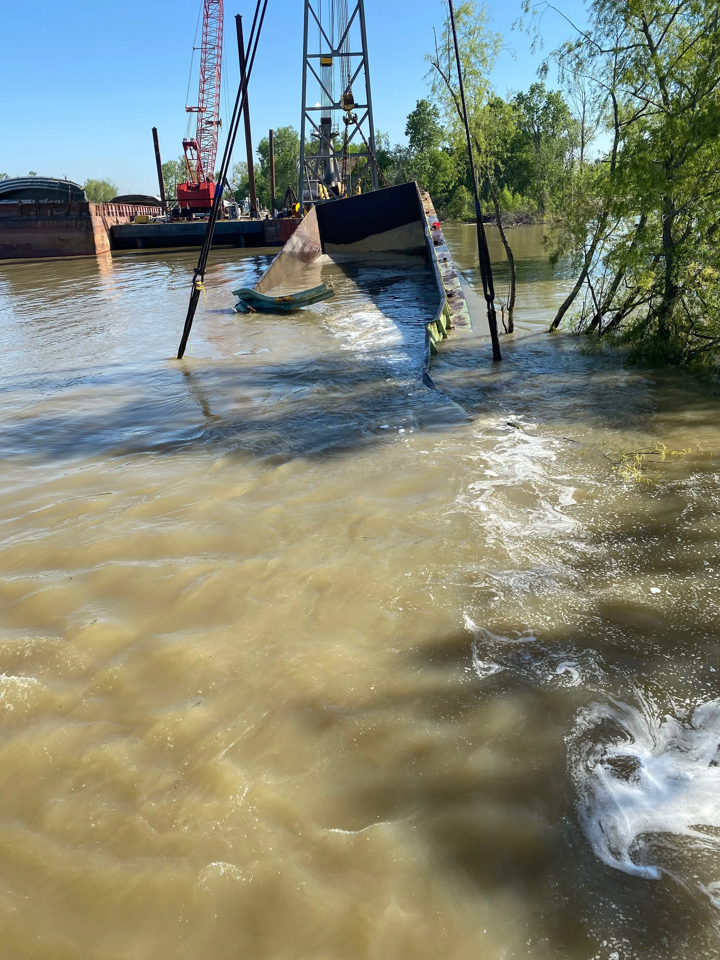 A partially submerged boat in a river, with construction cranes and equipment in the background, and trees on the riverbank.