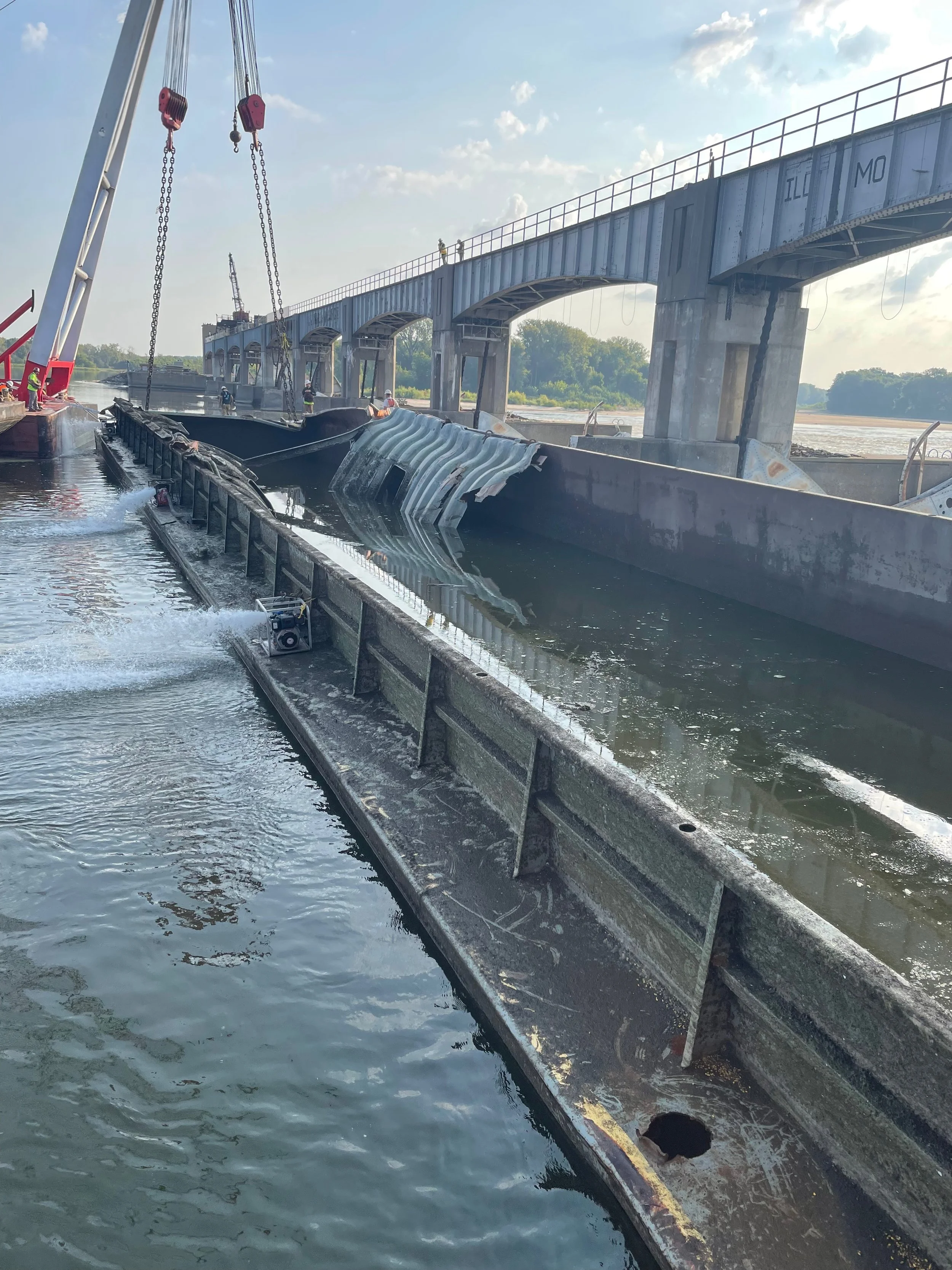 Construction workers load a large metal dam wall into the water using a crane, with a bridge and blue sky in the background.