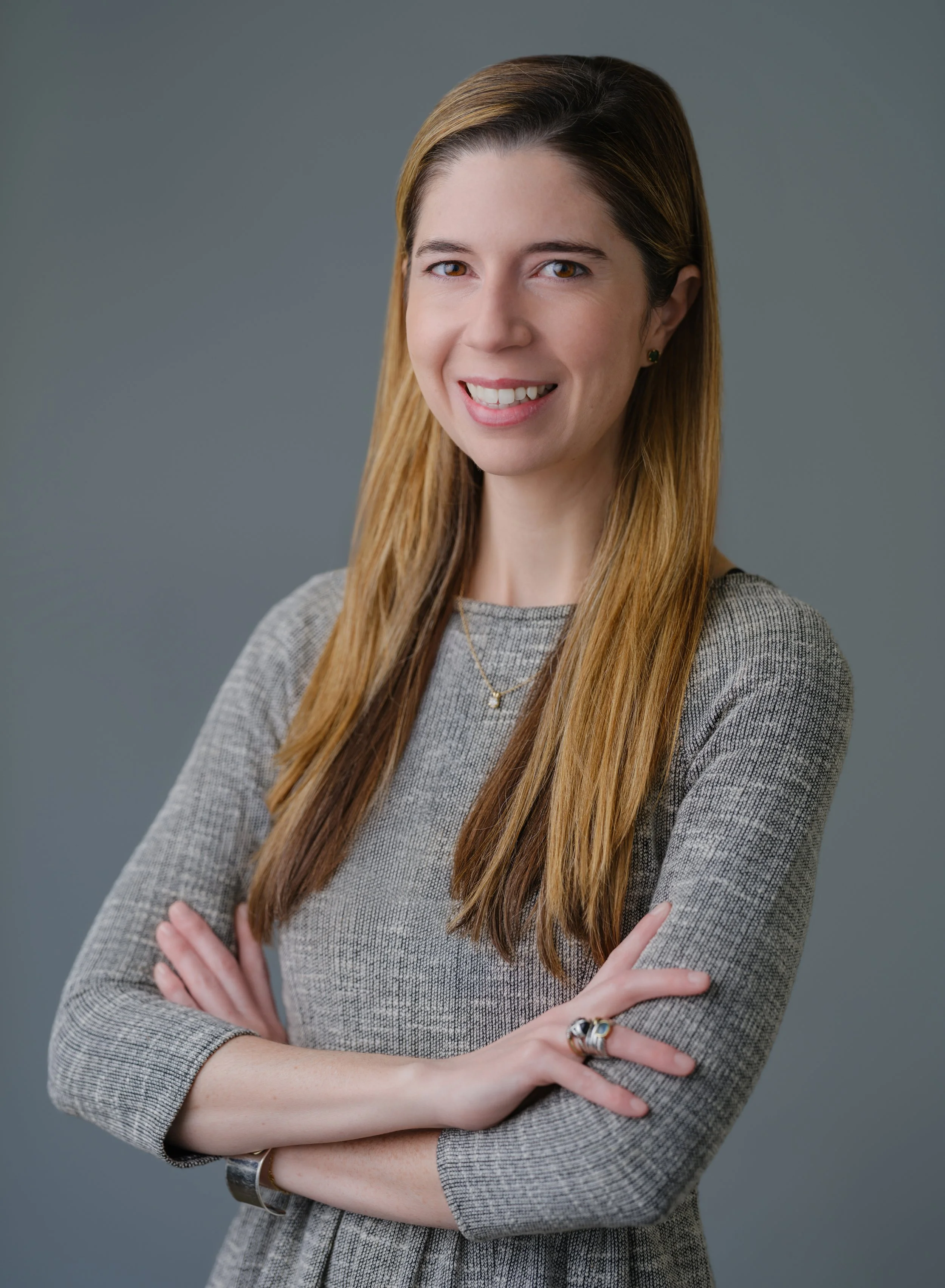 Portrait of a smiling woman with long, light brown hair, wearing a grey dress with her arms crossed, standing against a plain grey background.