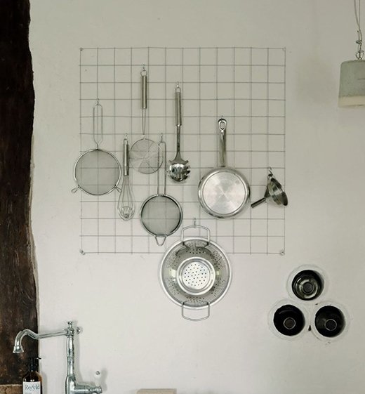 Kitchen wall with a grid displaying hanging metal kitchen utensils, including strainers, a whisk, a ladle, and a colander, along with three small black bowls on the right.