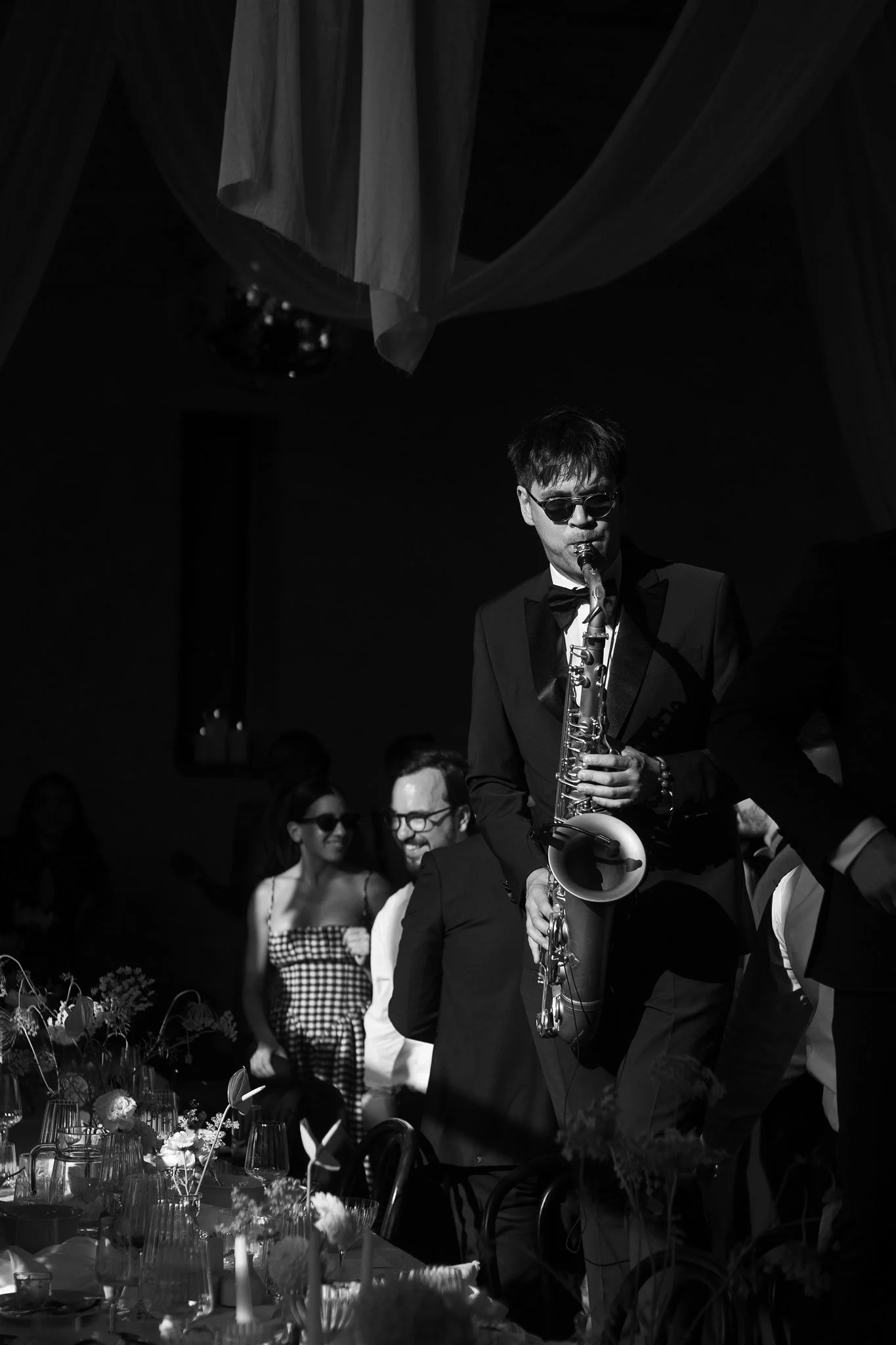 A black-and-white photo of a man playing the saxophone at a formal event. He is wearing sunglasses and a tuxedo, with a group of smiling people in the background, some wearing sunglasses as well. The setting appears to be a decorated indoor venue.