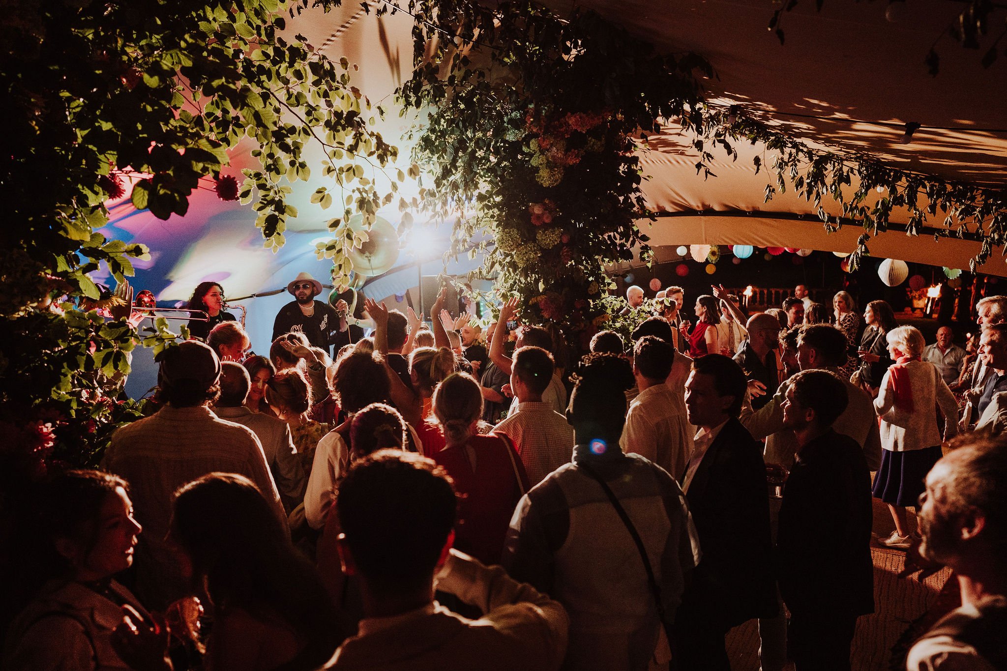 A lively indoor party scene with people dancing and socializing under a canopy decorated with greenery and flowers, with a band performing on stage in the background.