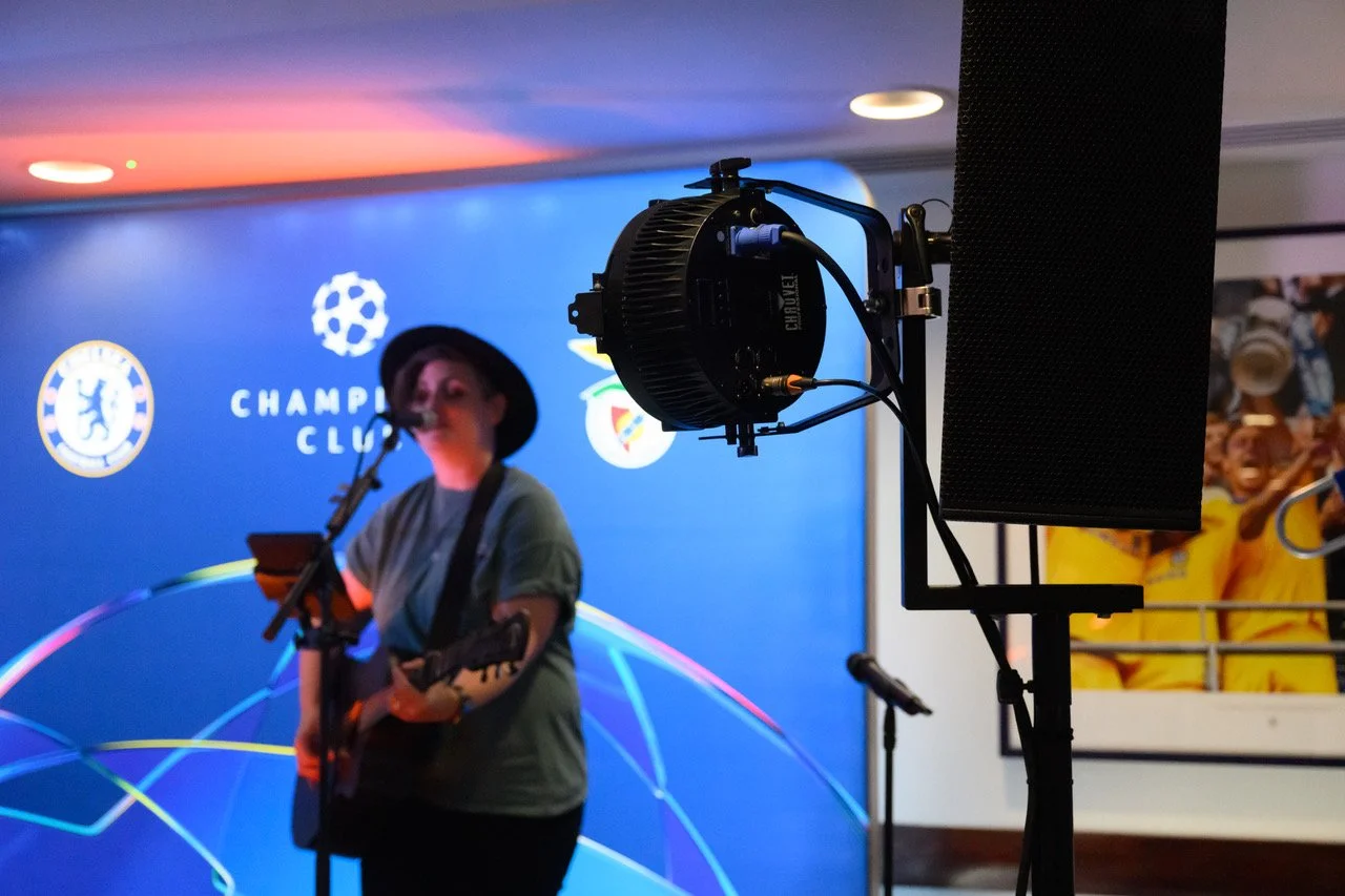 A woman wearing a hat and a green t-shirt is playing an acoustic guitar and singing into a microphone on stage at a Chelsea football club event. There is a speaker, stage lighting, and a blue background with the Chelsea logo and a football-themed des