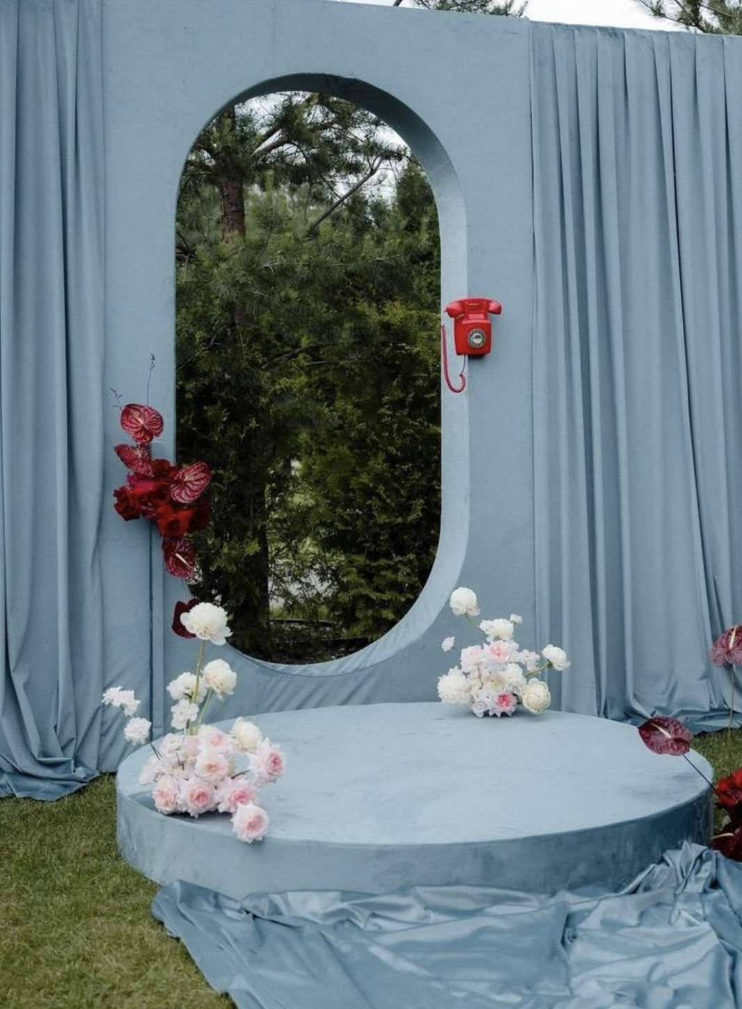 A stage with a blue curtain backdrop, a circular platform decorated with pink and white flowers, a red rotary phone mounted on the curtain, and a background of green trees visible through an oval opening.