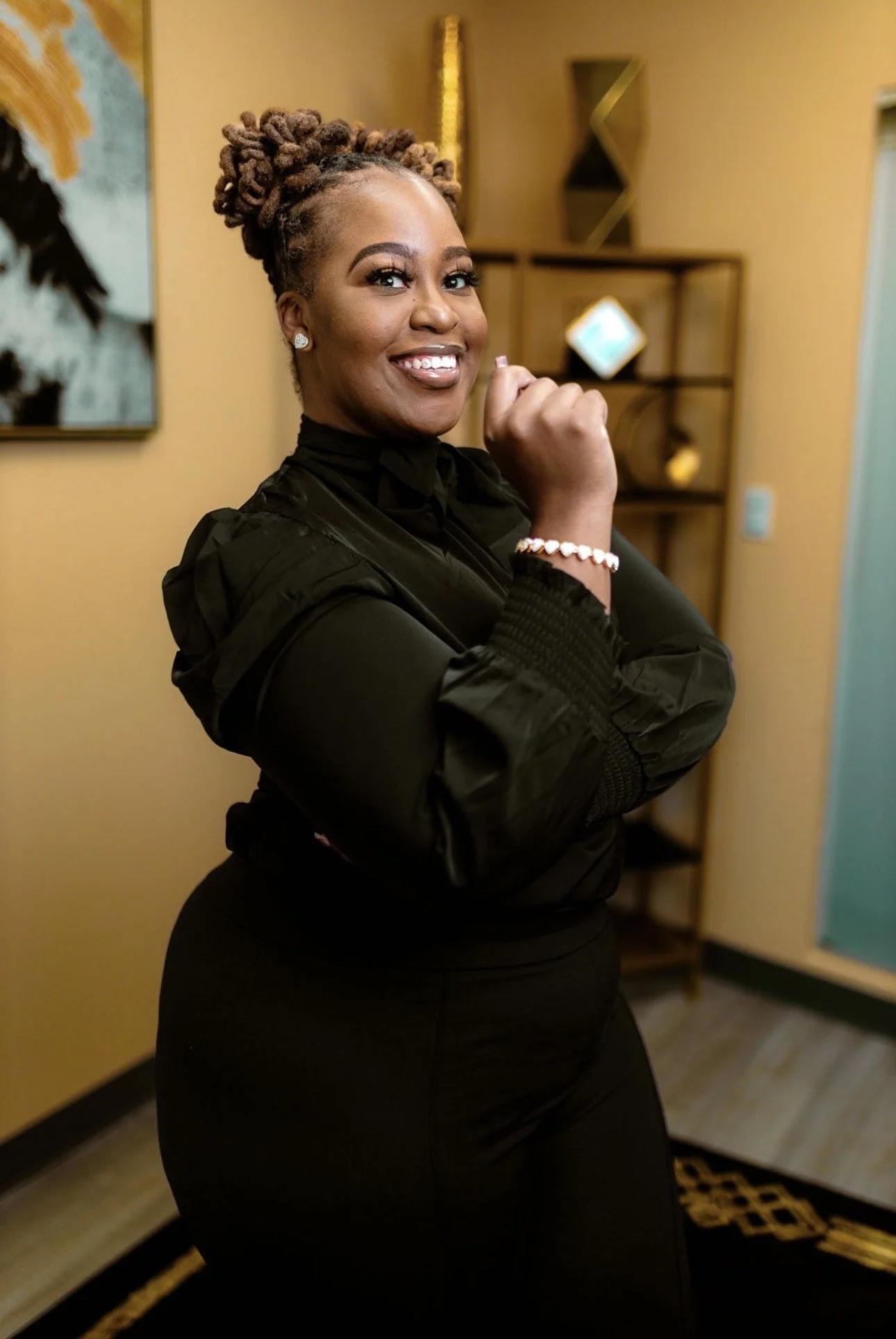 A smiling woman with styled hair, wearing a black outfit, jewelry, standing in a warmly decorated room.