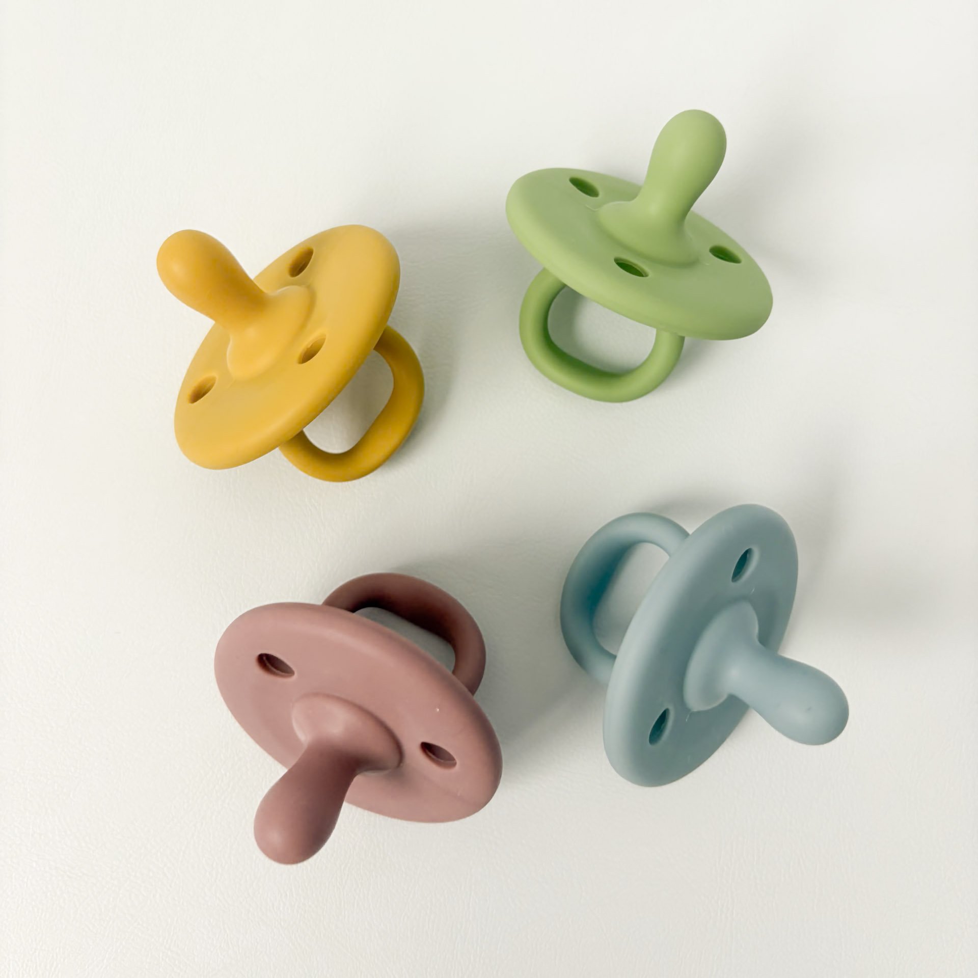Four colorful silicone pacifiers in yellow, green, pink, and blue, arranged on a white surface.