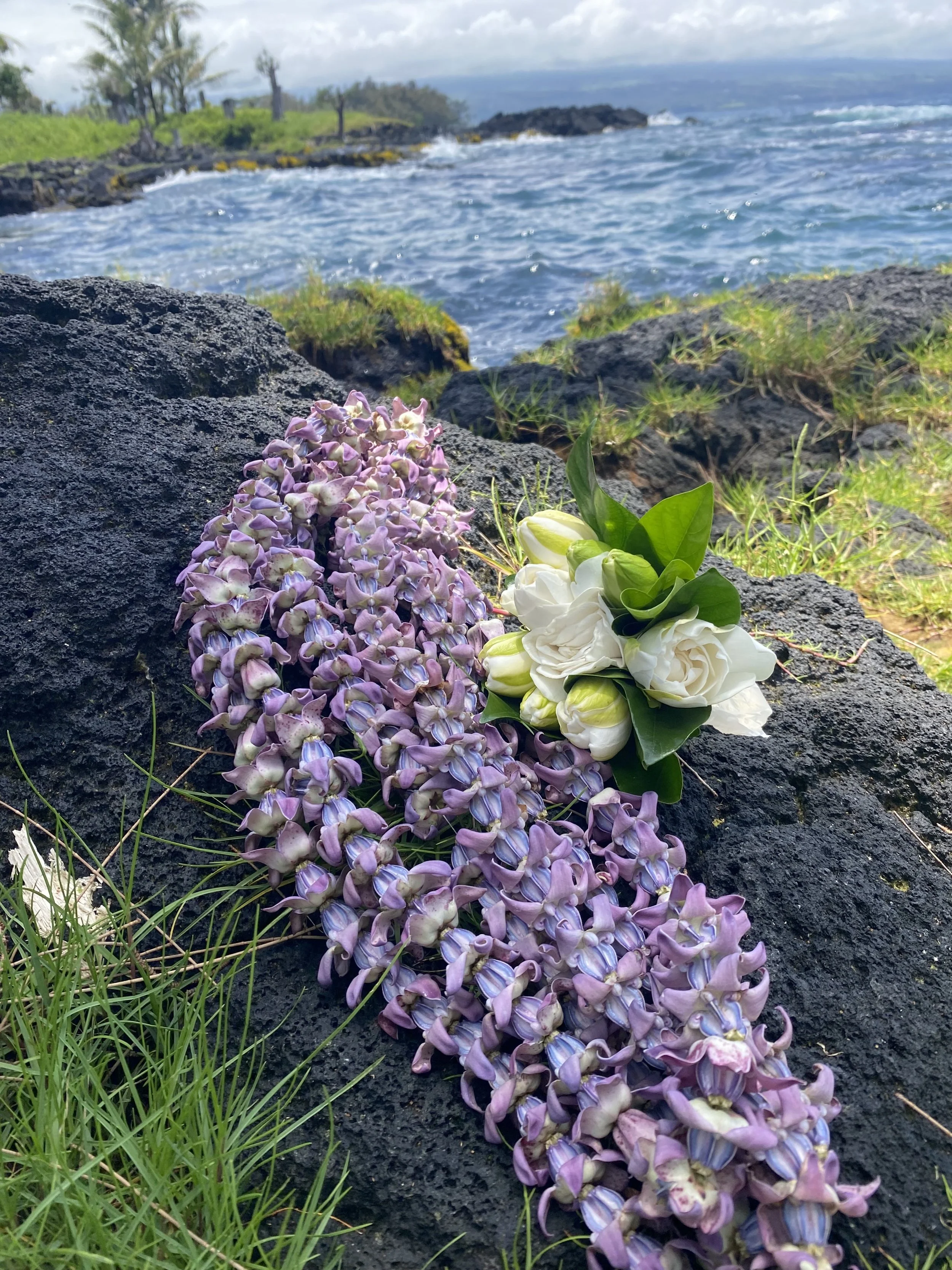 pua kalaunu lei and pikake flower resting on a rock infront of the ocean