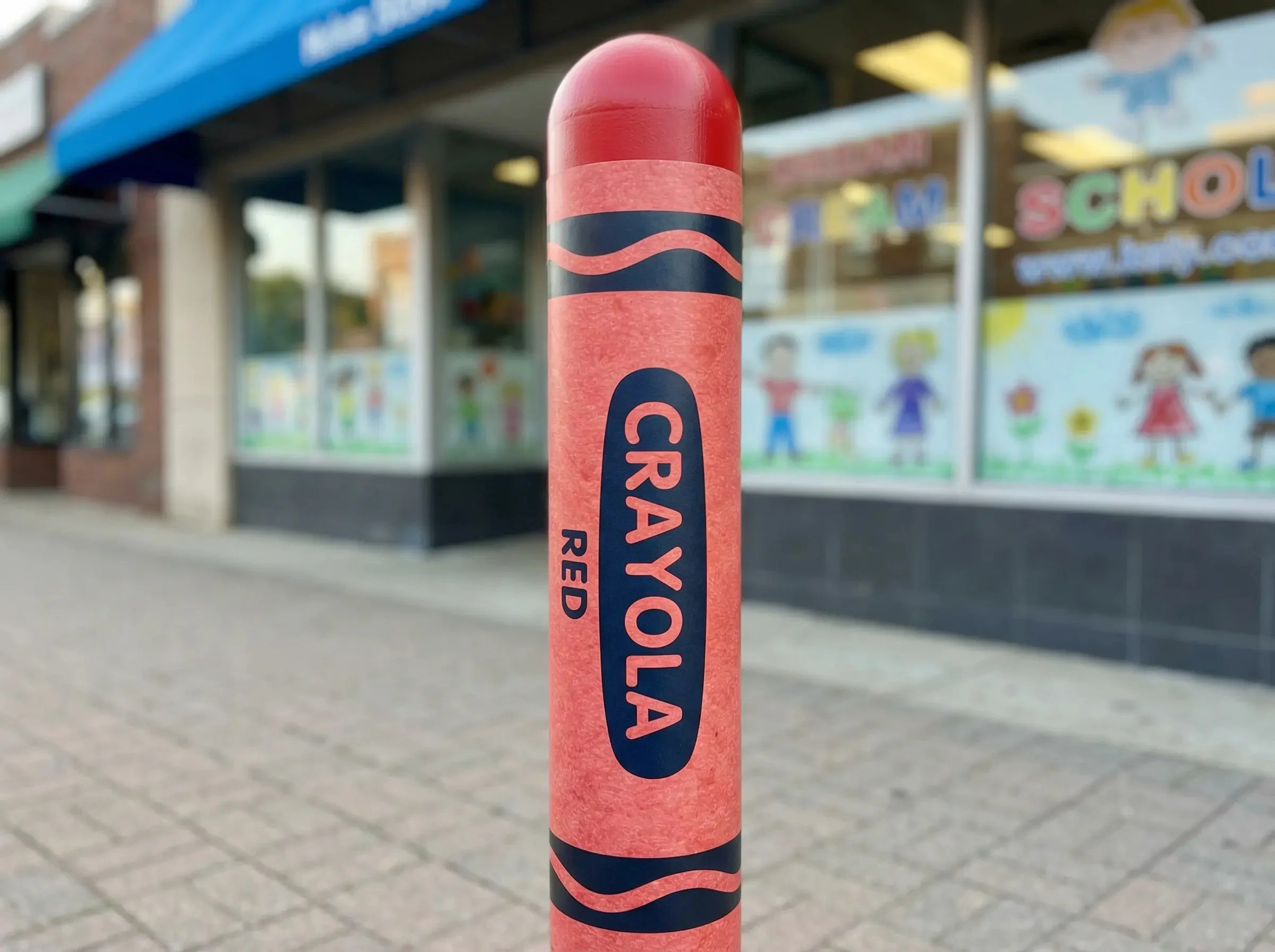 Pink pole with the words 'GAYOLA RED' printed on it, standing on a sidewalk outside a building with colorful children's artwork in the window.