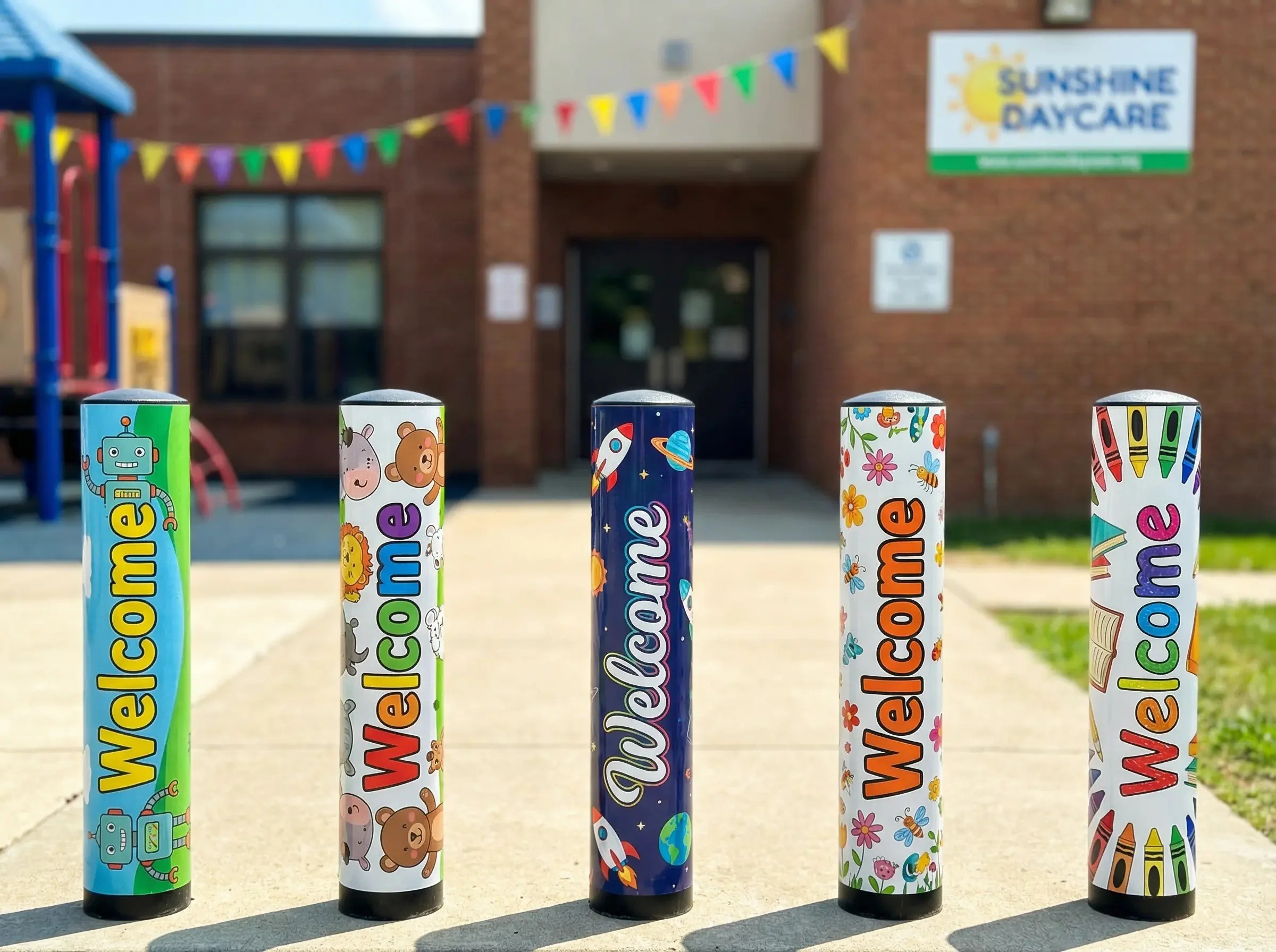 Five colorful cylindrical tubes with 'Welcome' written on each, decorated with various playful designs, on a concrete surface outside a building labeled Sunshine Daycare.
