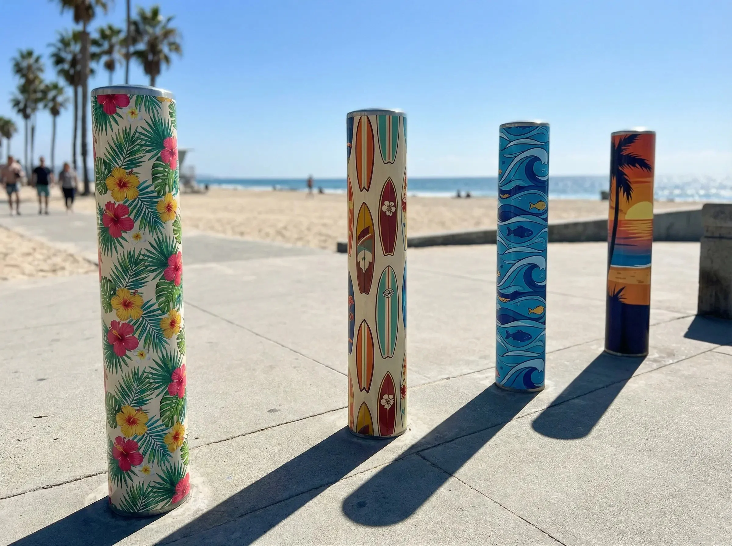 Four decorative cylindrical objects with colorful ocean and beach-themed patterns on a concrete surface near a beach, with palm trees, people, sand, and ocean in the background.