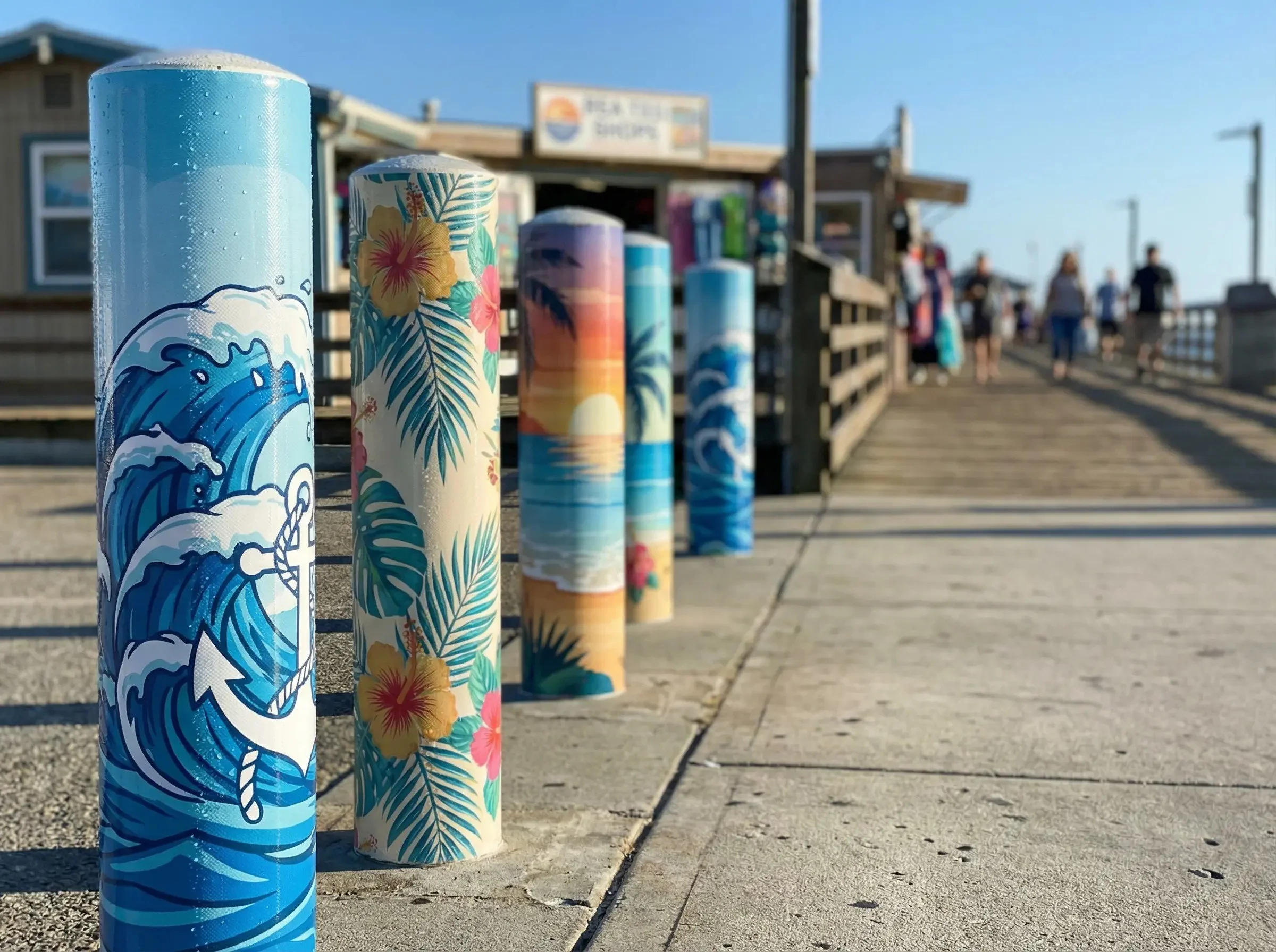 Colorful tiki-style poles with ocean and tropical designs line a pier walkway, with people walking in the background on a sunny day.