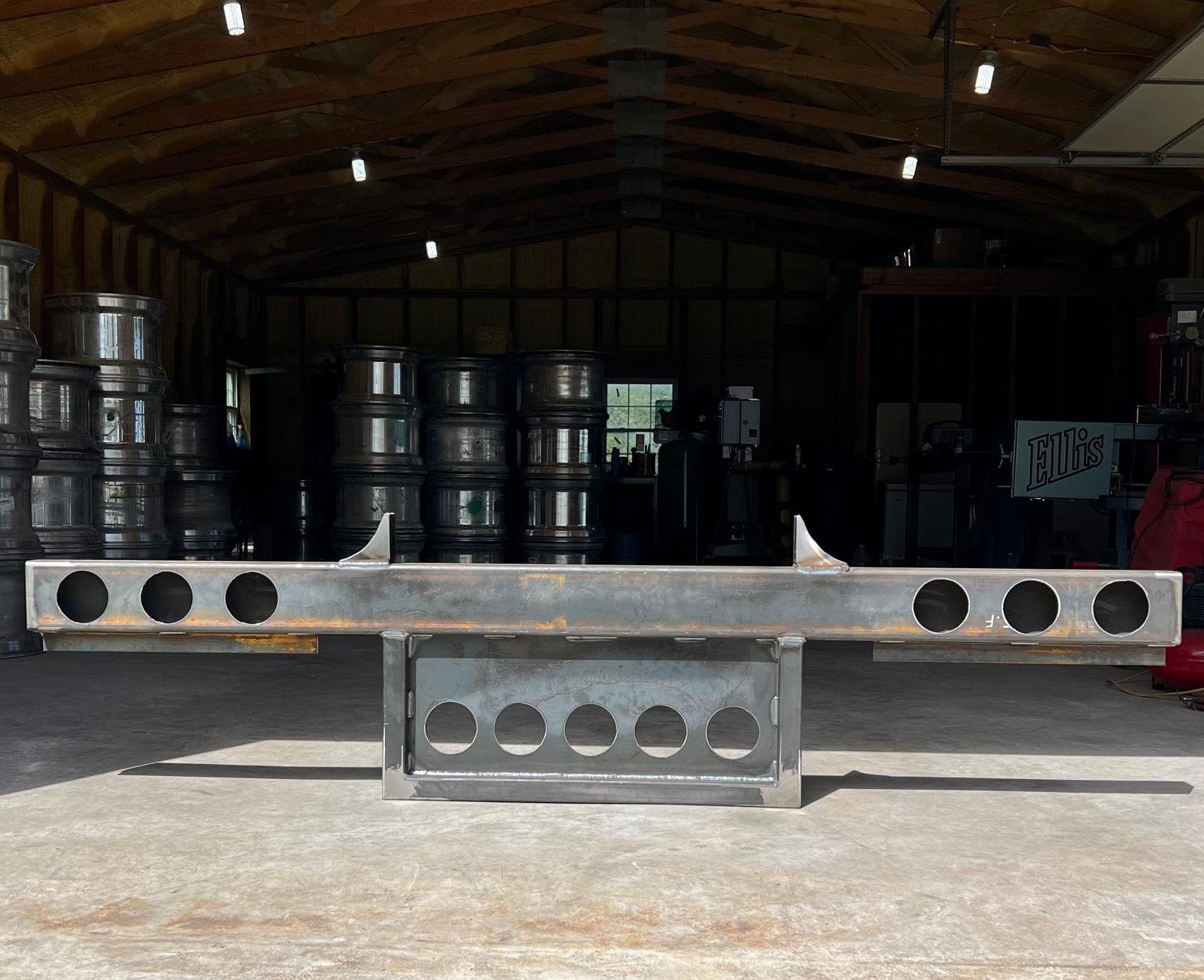 Metal machinery or tool with circular cutouts, positioned on a concrete floor inside a wooden workshop or garage, with stacked metal barrels and machinery in the background.