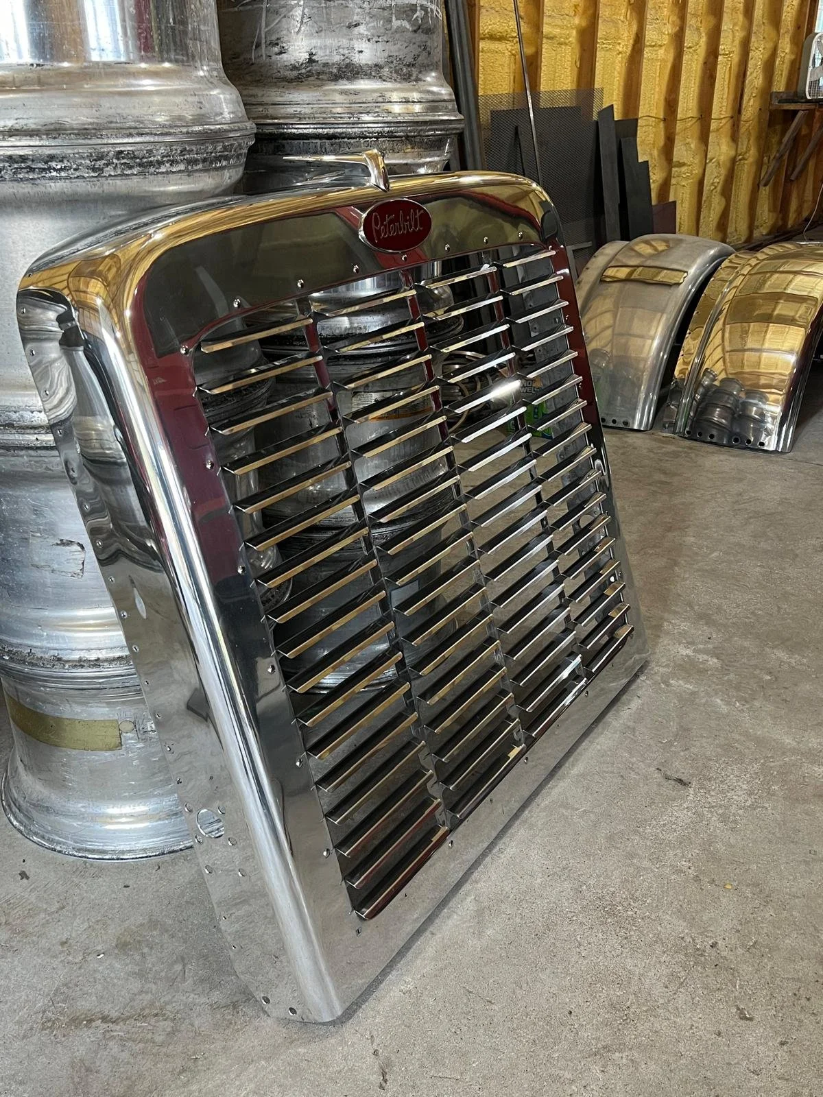 A Polished Metal truck grill with chrome finish, leaning against a shelf with metal wheel rims and car fenders in a workshop or garage.