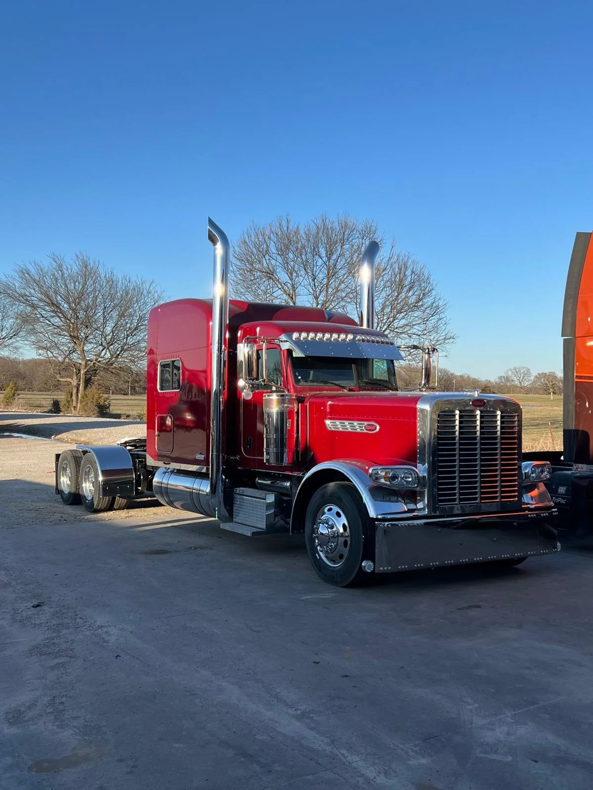 Red semi-truck parked outdoors on a paved surface with leafless trees in the background and a clear blue sky.