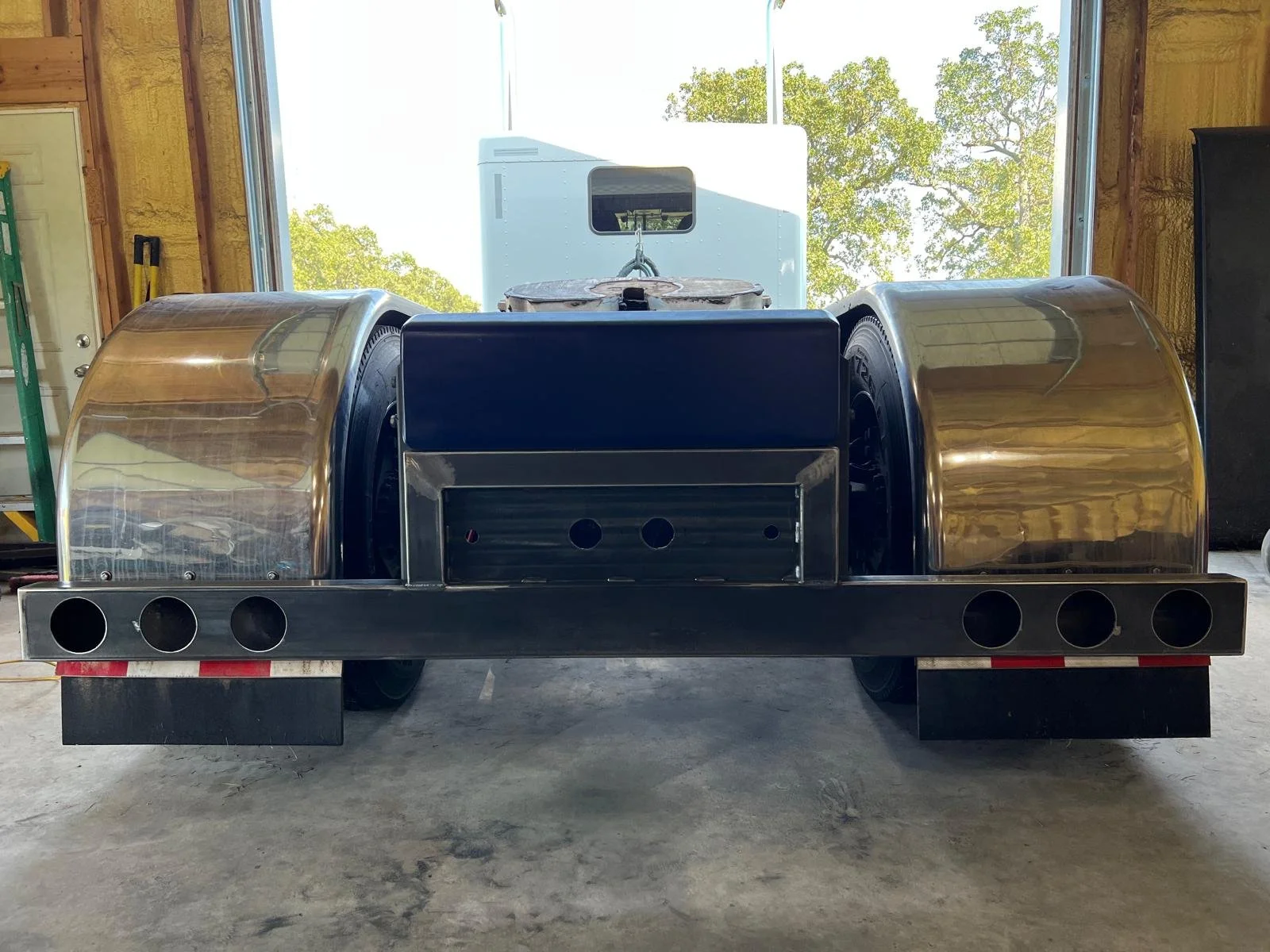 Back view of polished semi-truck fenders inside a workshop, with a trailer and trees visible through an open garage door in the background.
