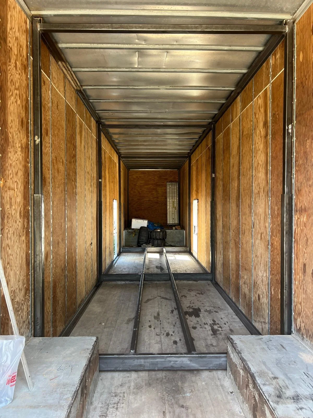 Interior of a partially constructed cargo trailer with wooden walls, metal framework, and tracks on the floor for securing items, with construction tools and materials in the back.