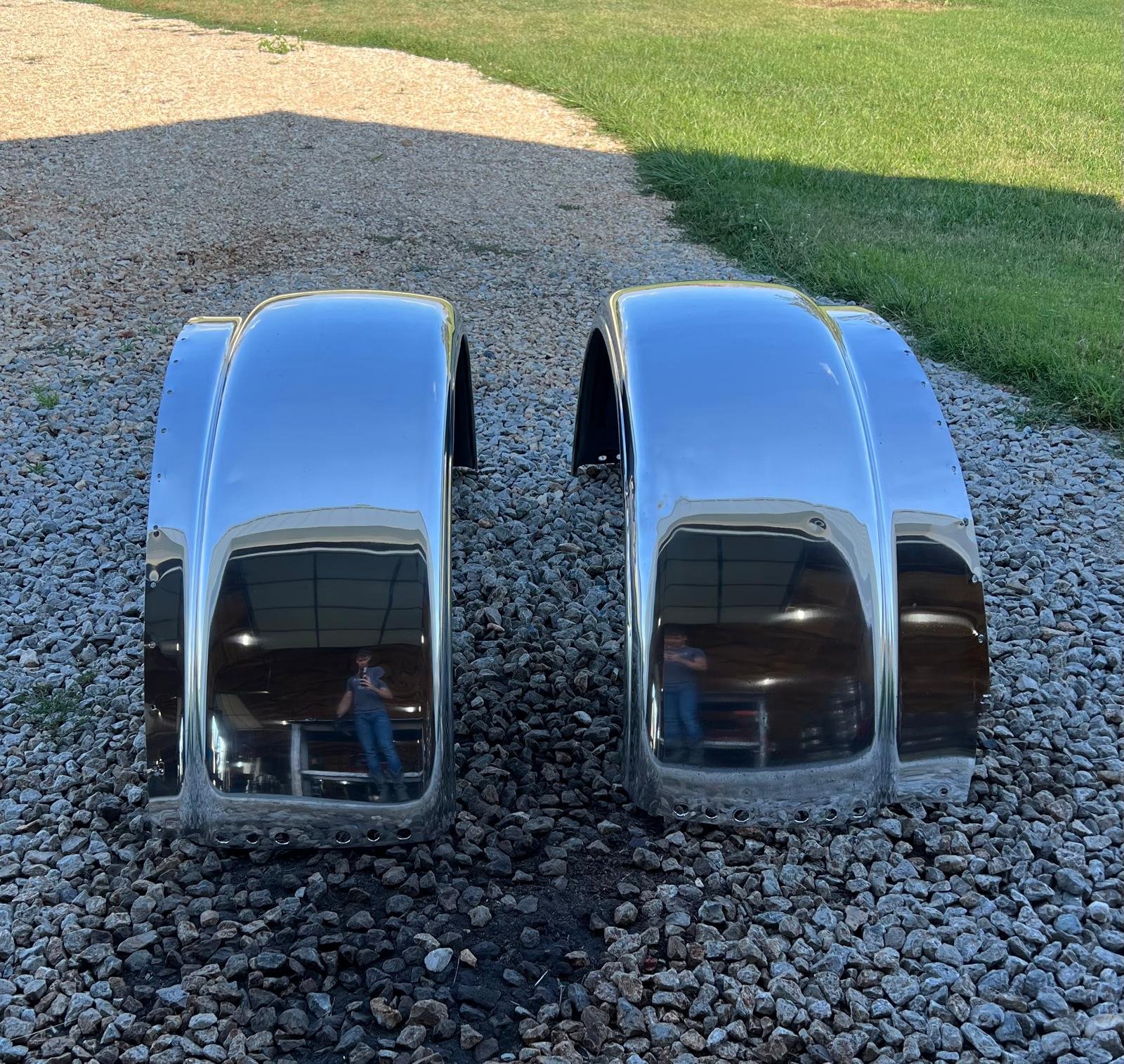 Two polished chrome truck fenders, placed on a gravel surface outside. Their reflective surfaces mirror the sky, surrounding environment, and the person taking the photo.