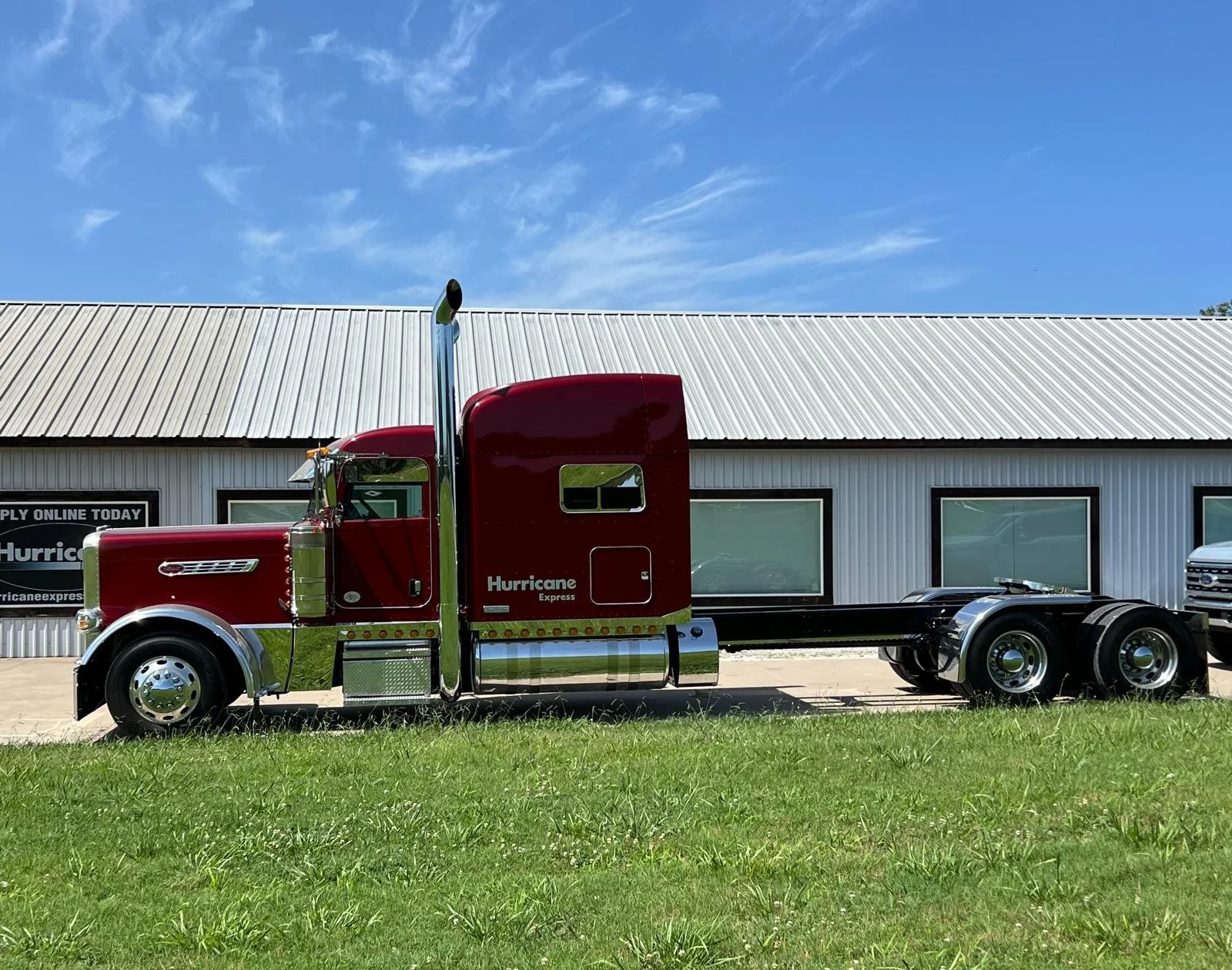 A red semi-truck with the name 'Hurricane Express' on the side, parked in front of a white metal building with a metal roof, under a blue sky with some clouds.