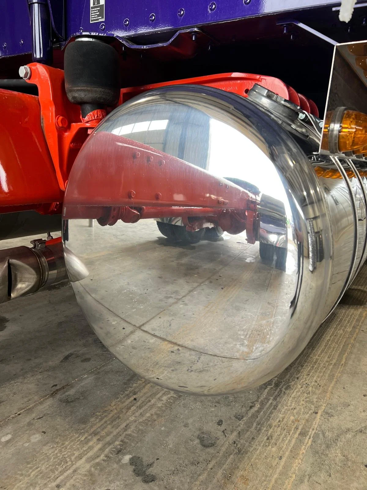 Close-up of a shiny, chrome motorcycle fuel tank with a reflection of a garage or warehouse interior in it. The tank is mounted on a red motorcycle frame, with a black saddle and part of the exhaust visible.