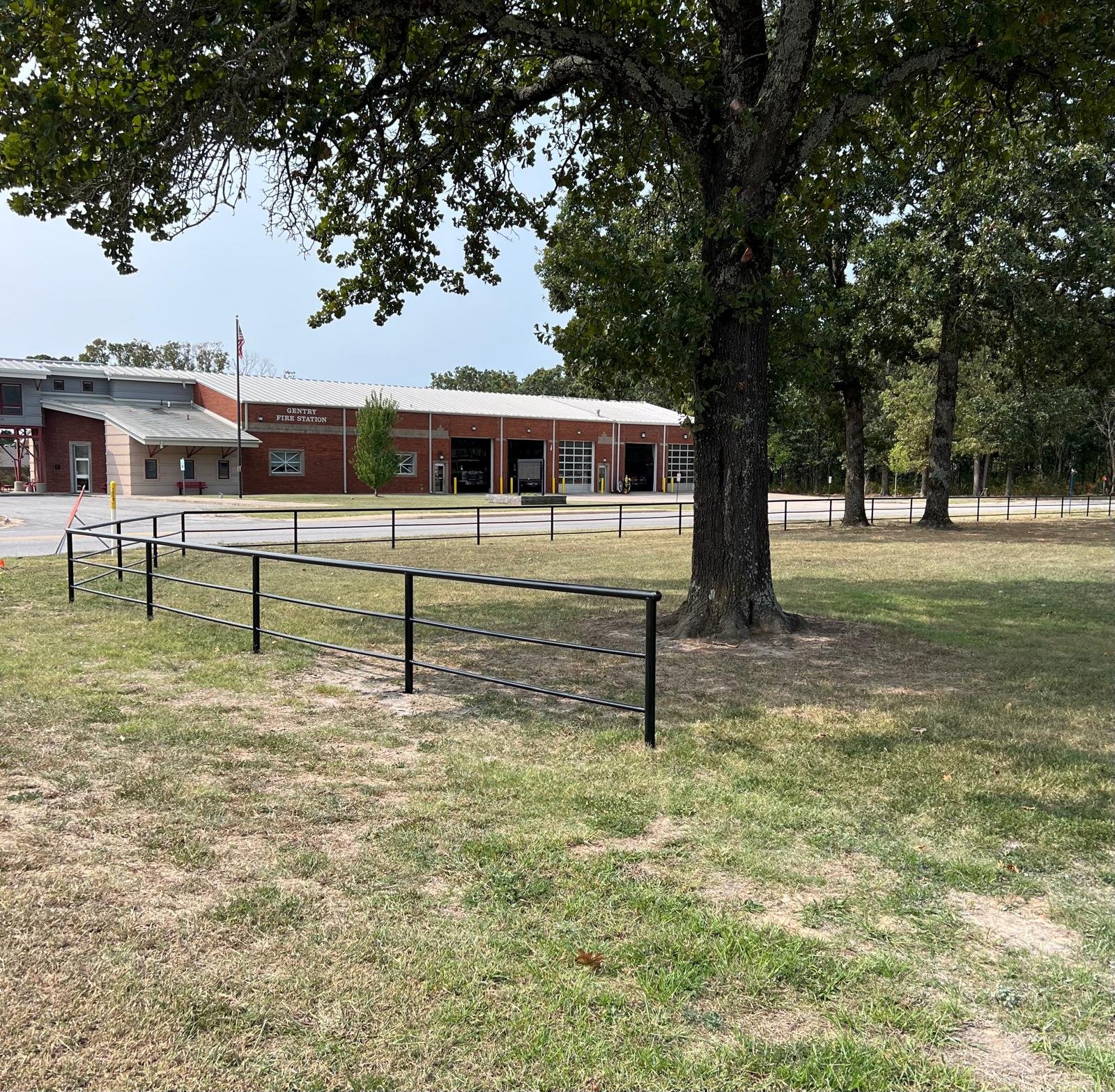 A fire station building with a red brick exterior, garage bays, and an American flag flying in front, surrounded by trees and a grassy area with a black metal fence.