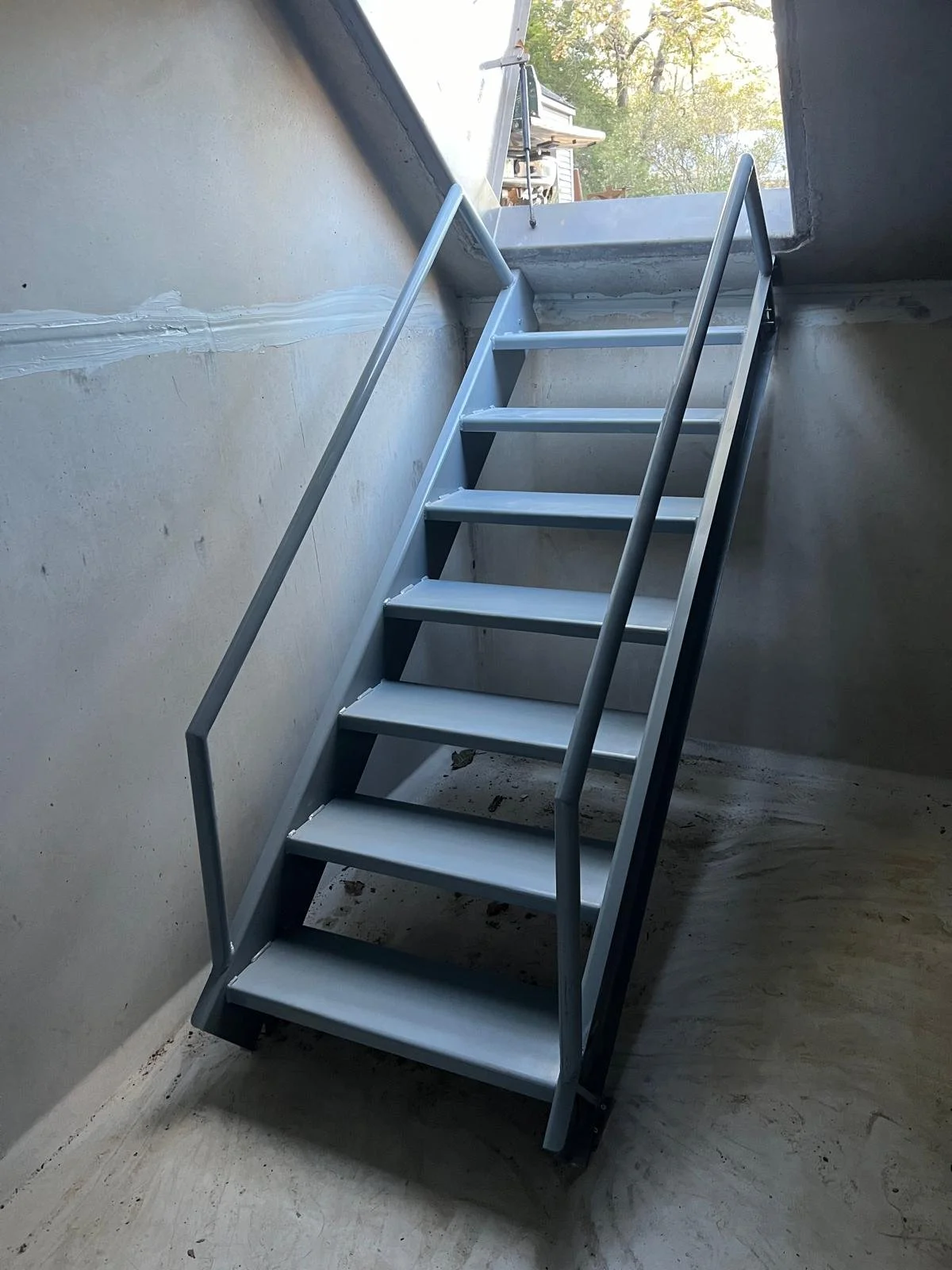 Metal staircase leading up through an open hatch in the ceiling, with a view of trees and sunlight outside.