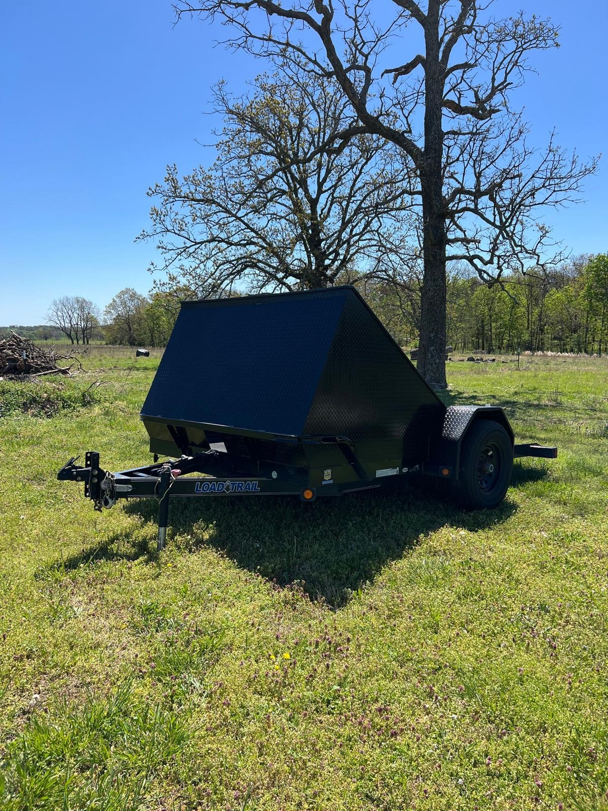 A black utility trailer with a tilted top is parked on a grassy field under a clear blue sky. There is a large tree without leaves behind the trailer and a smaller tree and open countryside in the distance.