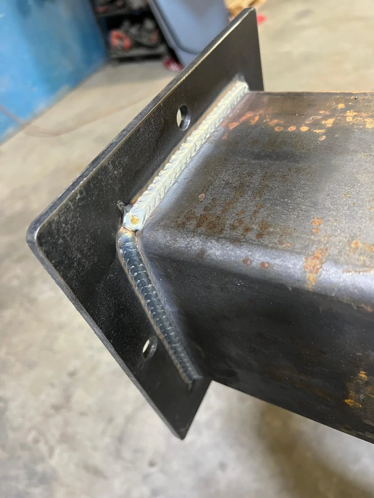 Close-up of a metal structure showing a welded joint attaching a piece of black painted metal to a rusted, greasy metal surface. The background is a workshop or garage setting.