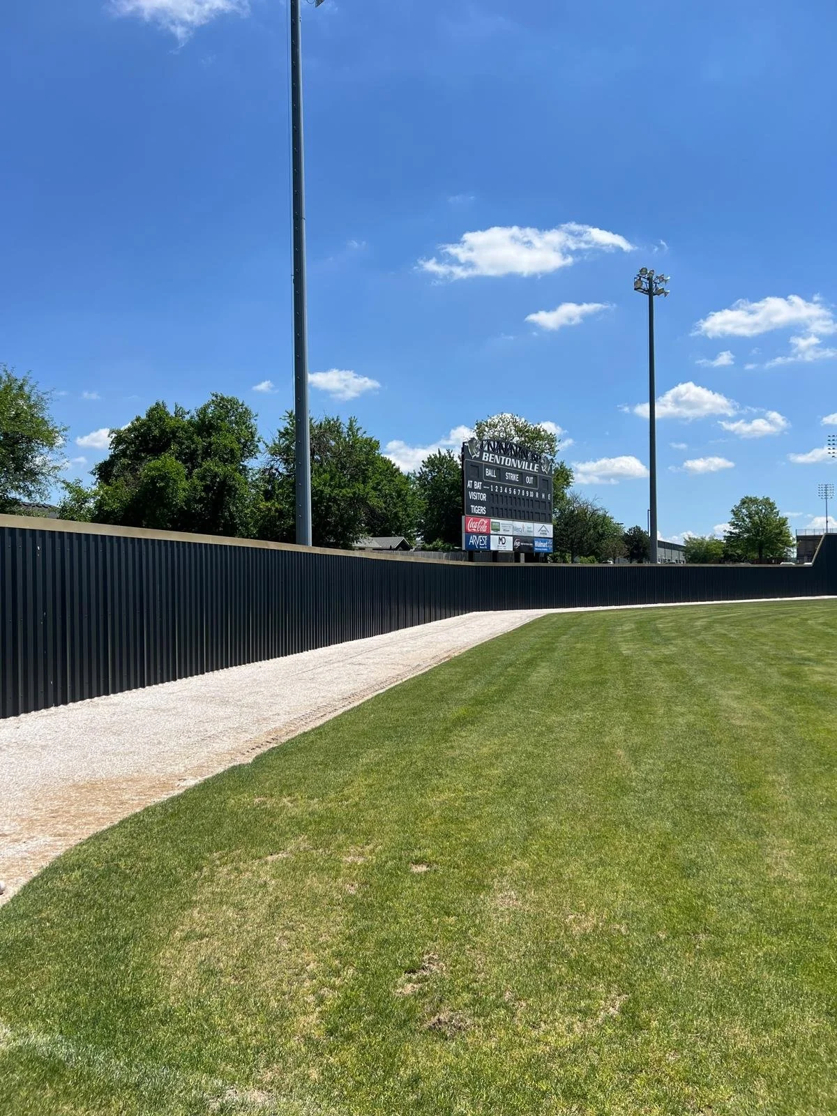 View of a baseball field with a scoreboard, green grass, a black fence, and tall light poles under a blue sky with some clouds.