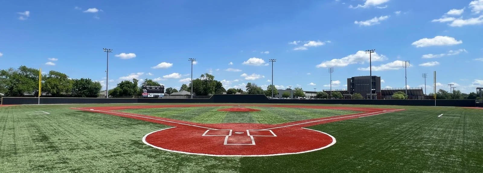 Empty baseball field under a clear blue sky with scattered clouds, surrounded by green trees and light poles.