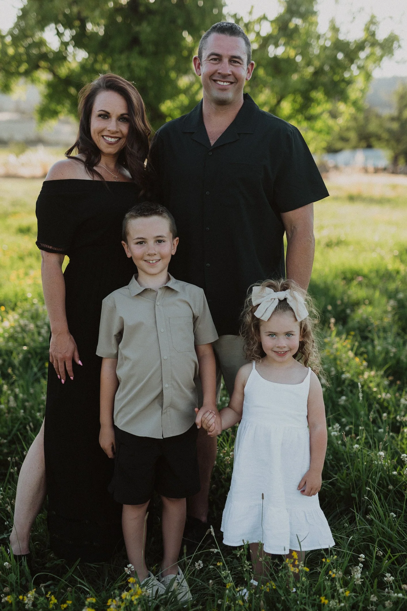A family of five standing outdoors in a grassy area with trees, smiling at the camera. The mother wears a black off-shoulder dress, the father in a black shirt, the boy in a beige shirt and shorts, and the girl in a white dress with a bow in her hair.