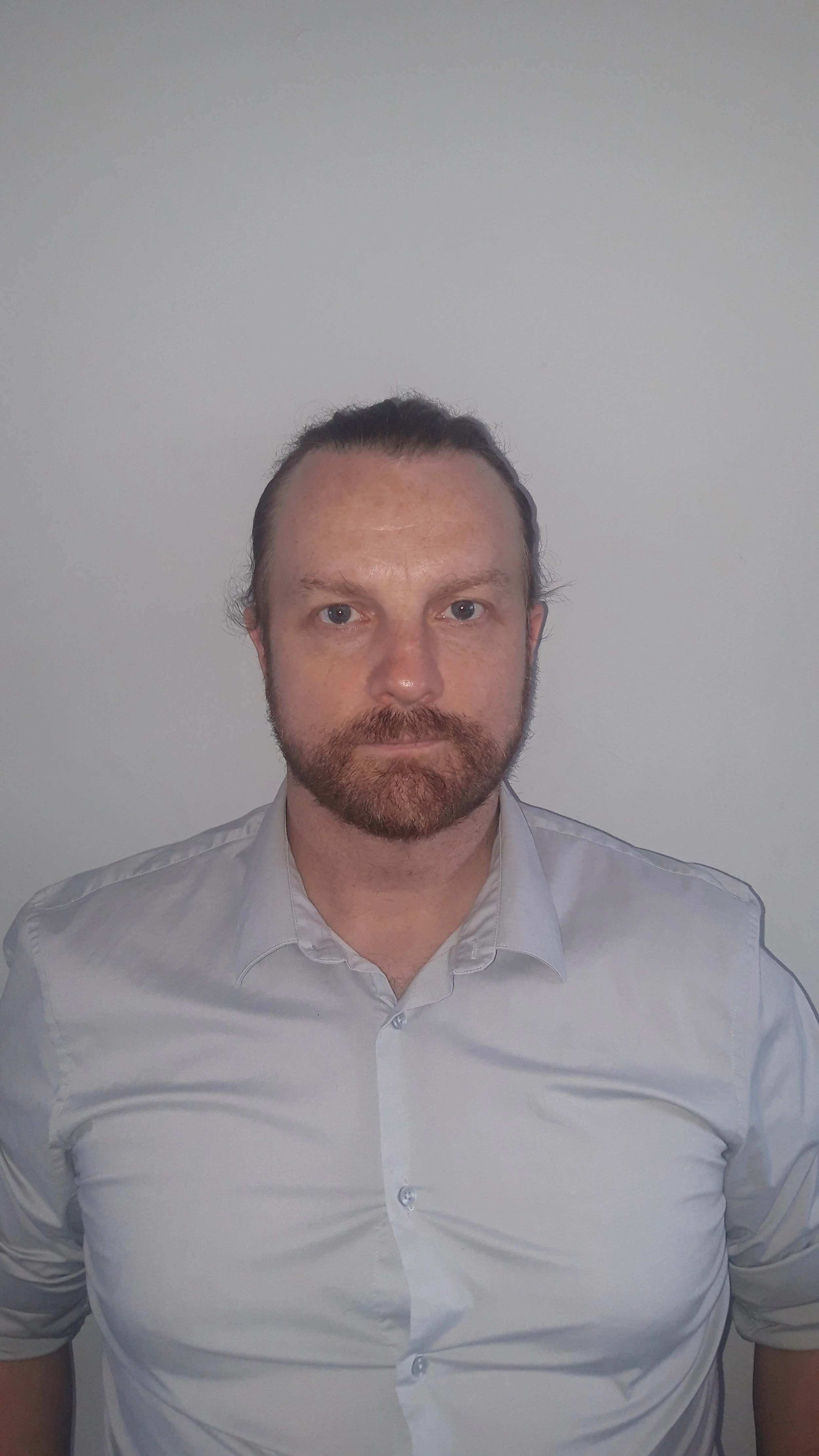 Portrait of a man with light skin, dark hair pulled back, a beard, wearing a light-colored button-up shirt, standing against a plain light-colored wall.