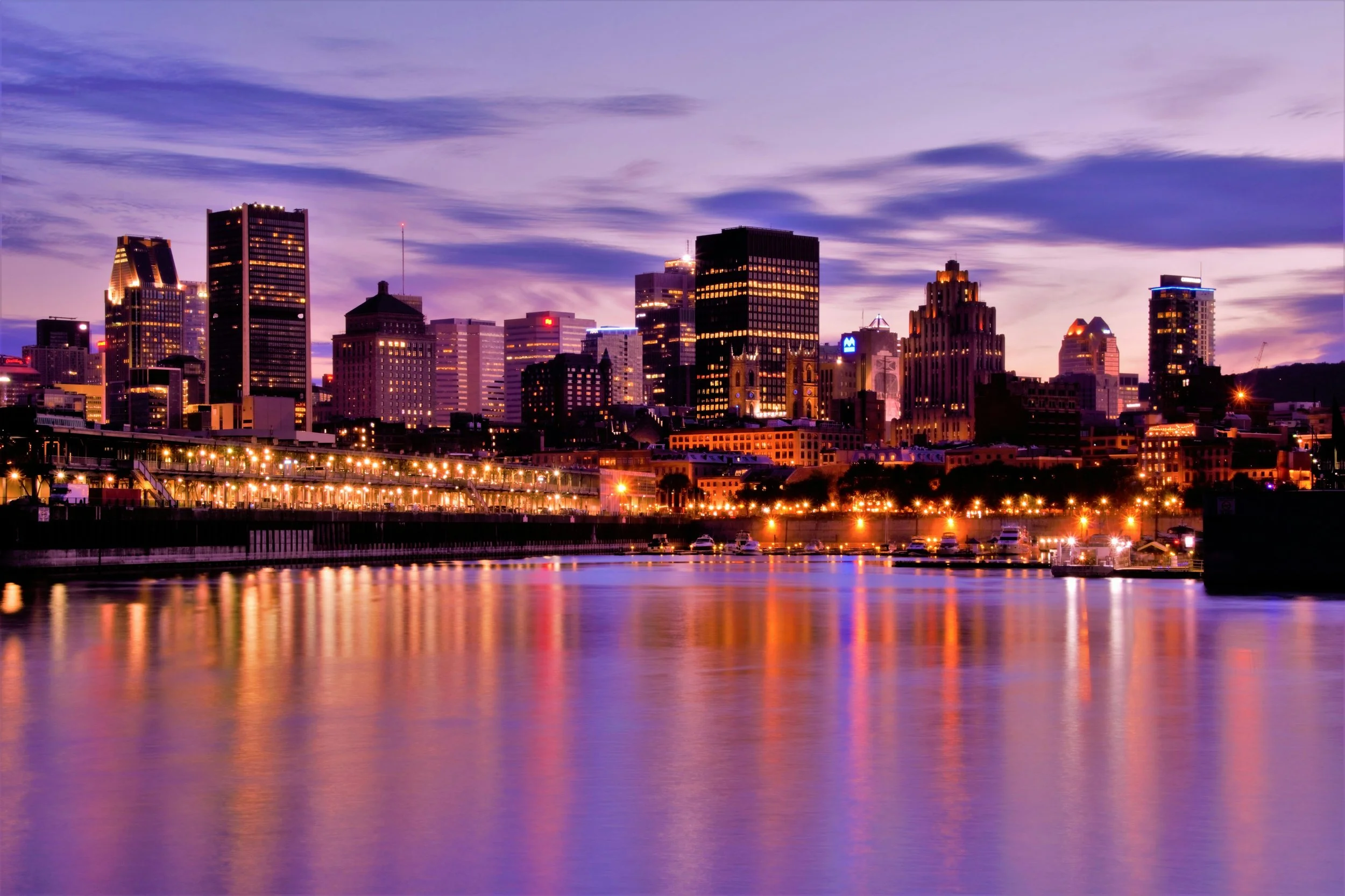 City skyline at dusk with illuminated buildings and reflections on the water.