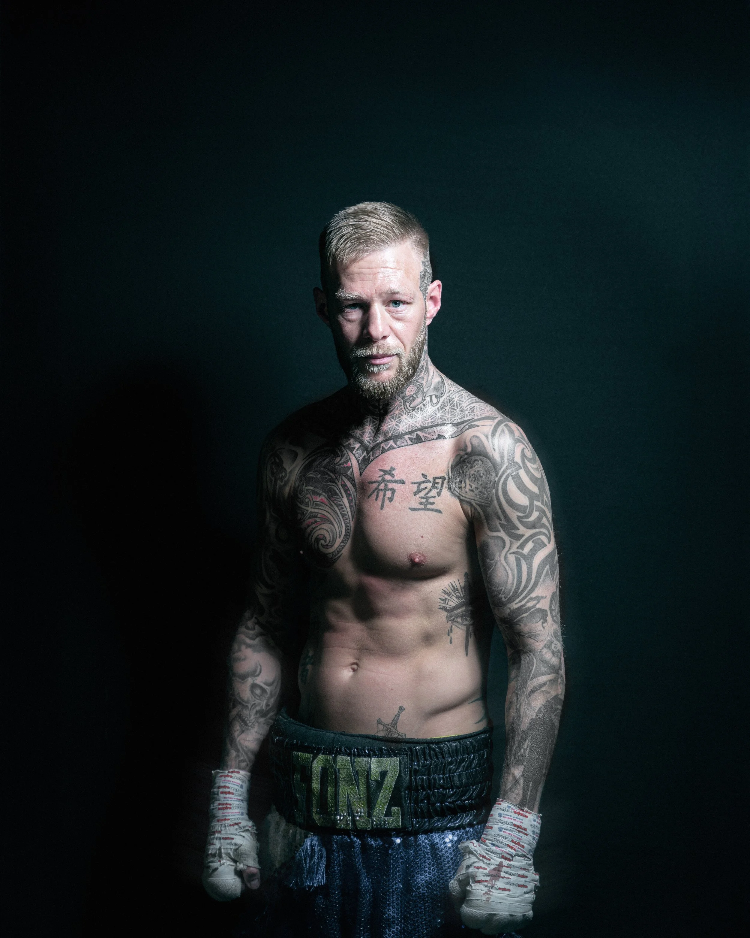 A shirtless man with tattoos standing against a dark background.