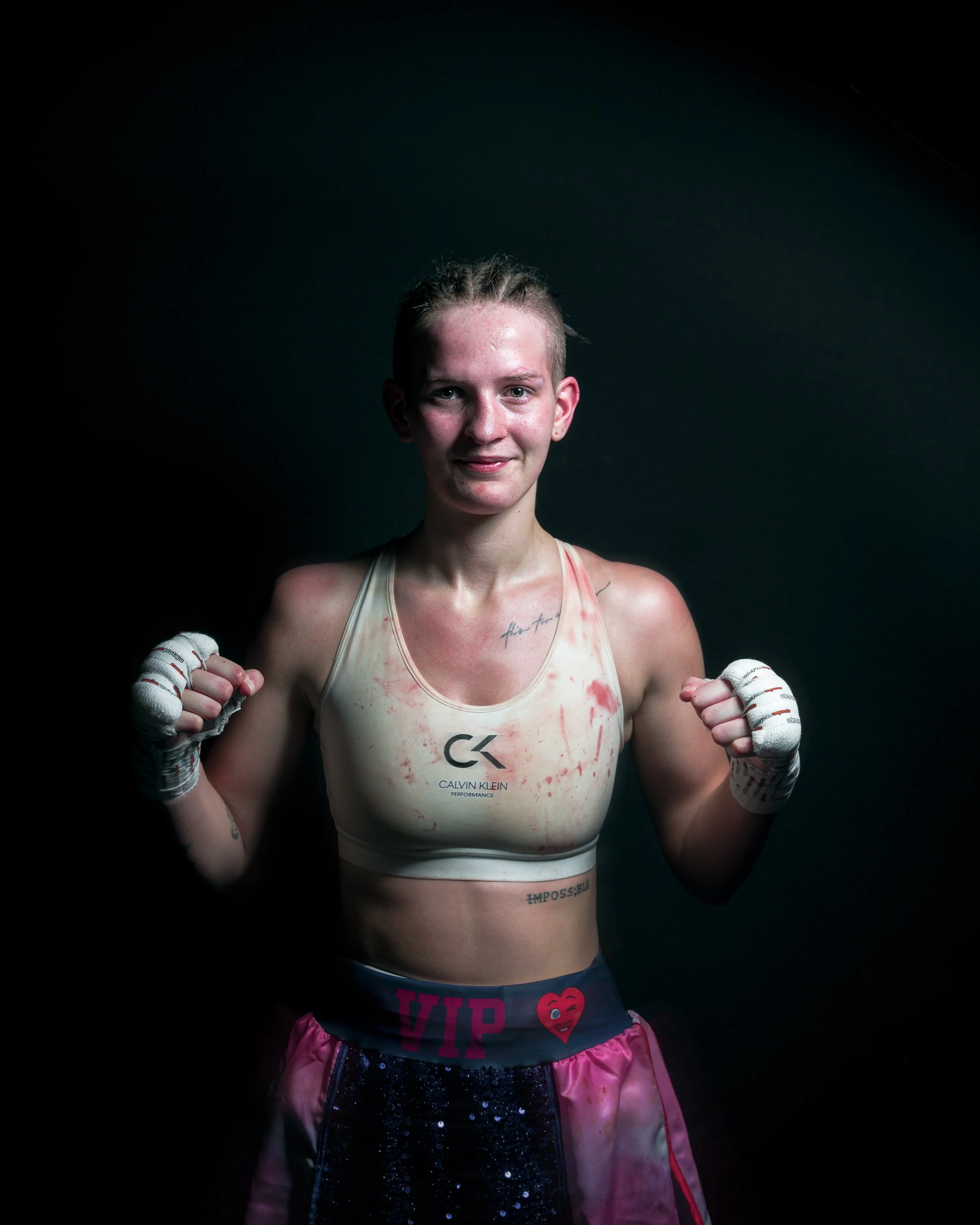 Female boxer in sports bra and shorts, smiling with fists raised, wearing hand wraps, with bloodstains on her top, standing against a dark background.