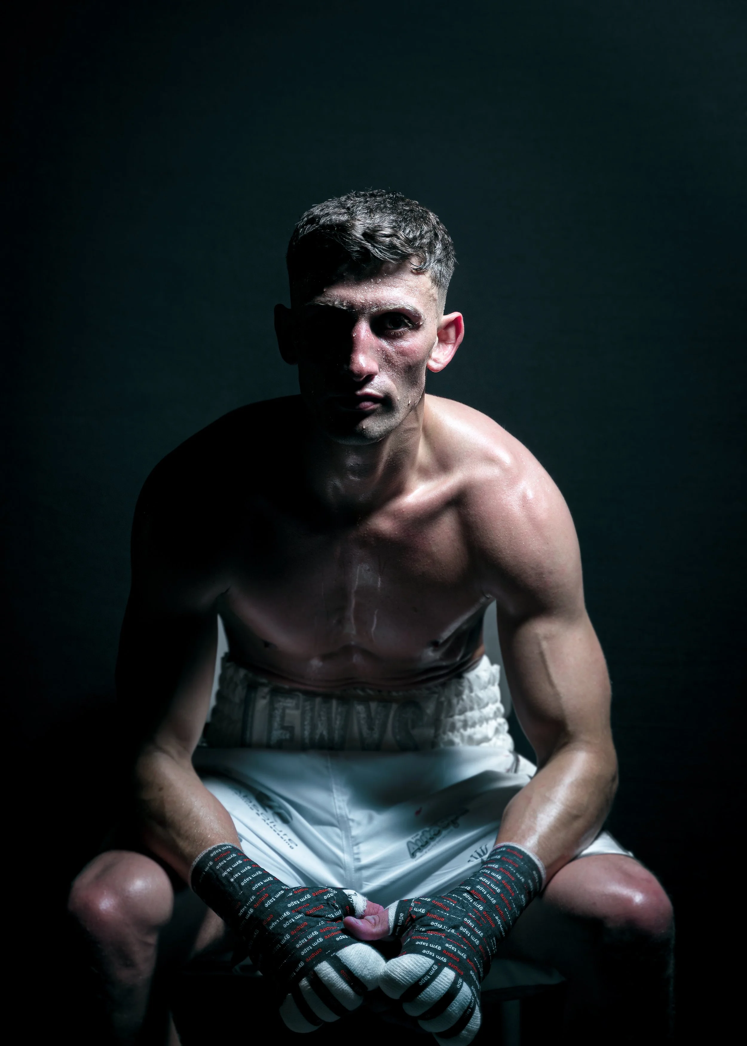 A shirtless male boxer sitting on a stool wearing boxing gloves, with intense lighting creating shadows on his face and body, against a dark background.