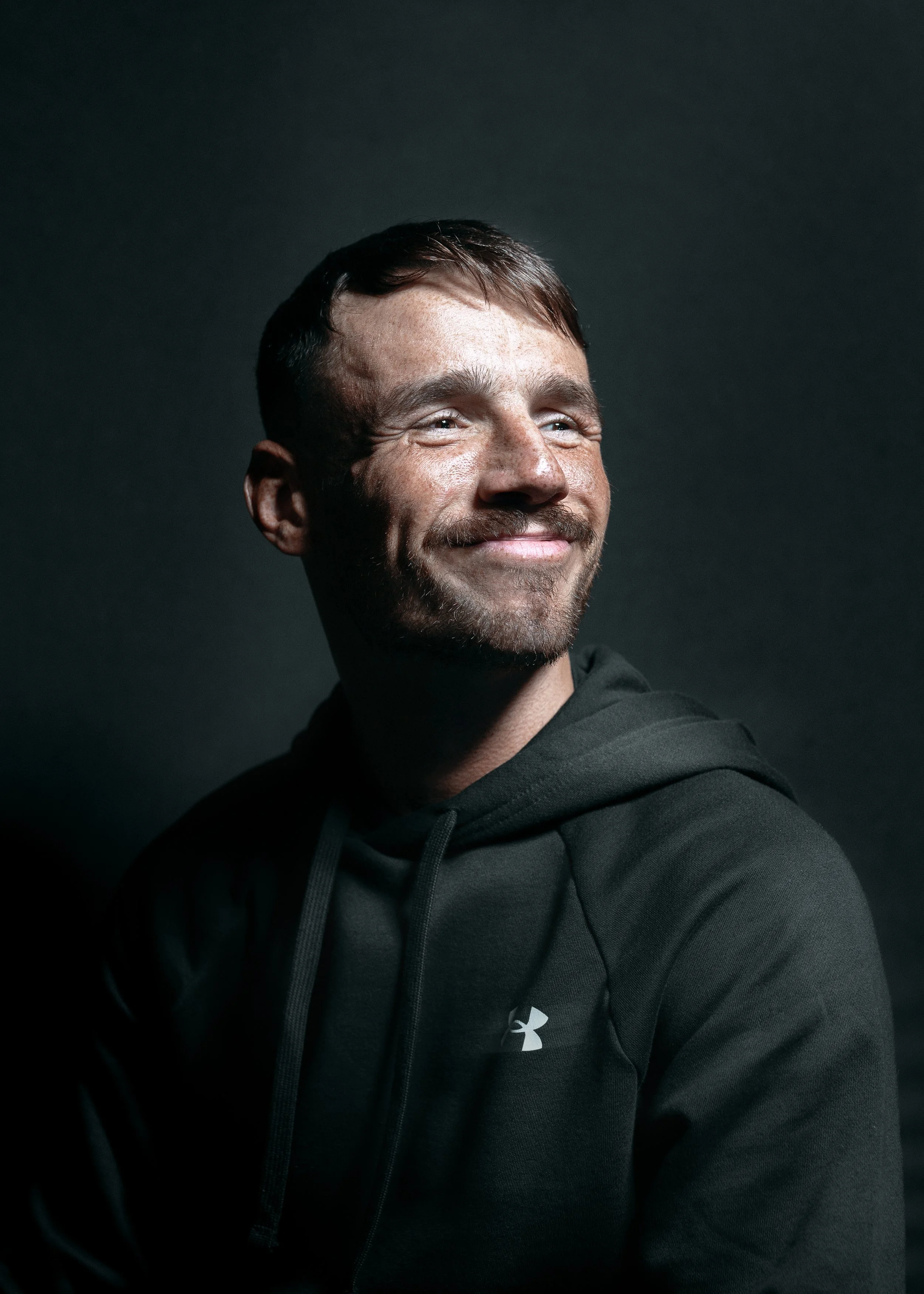 A man with short brown hair and a beard smiling while wearing a black Under Armour hoodie, set against a dark background.