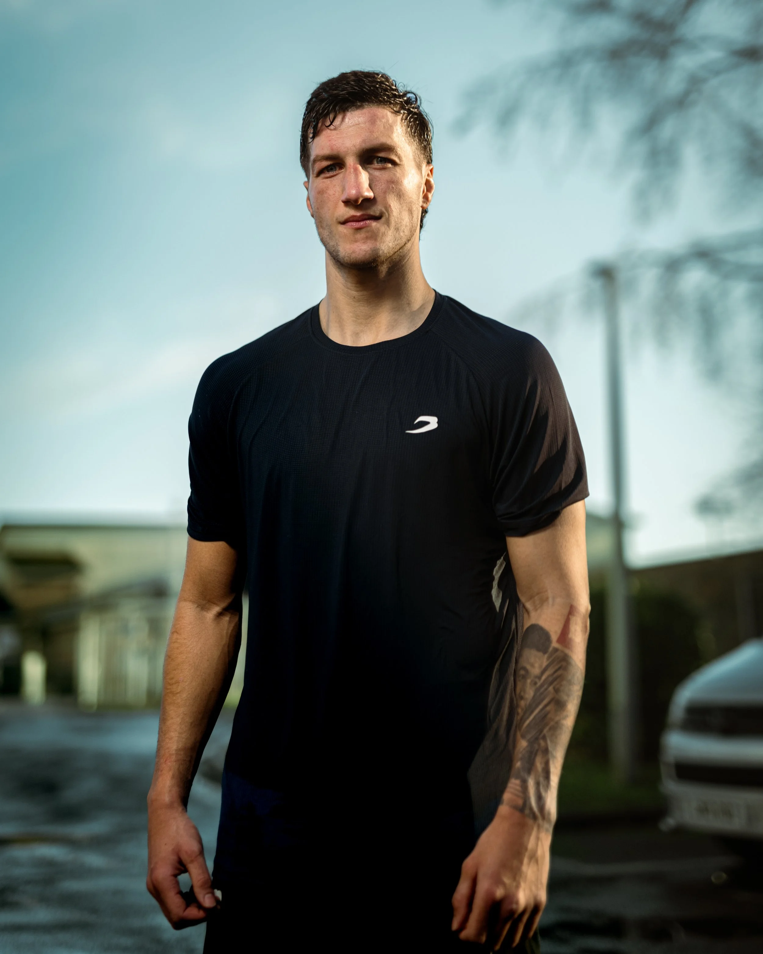 A young man standing outdoors wearing a black athletic shirt with a white logo, with tattoos on his left arm, on a cloudy day.