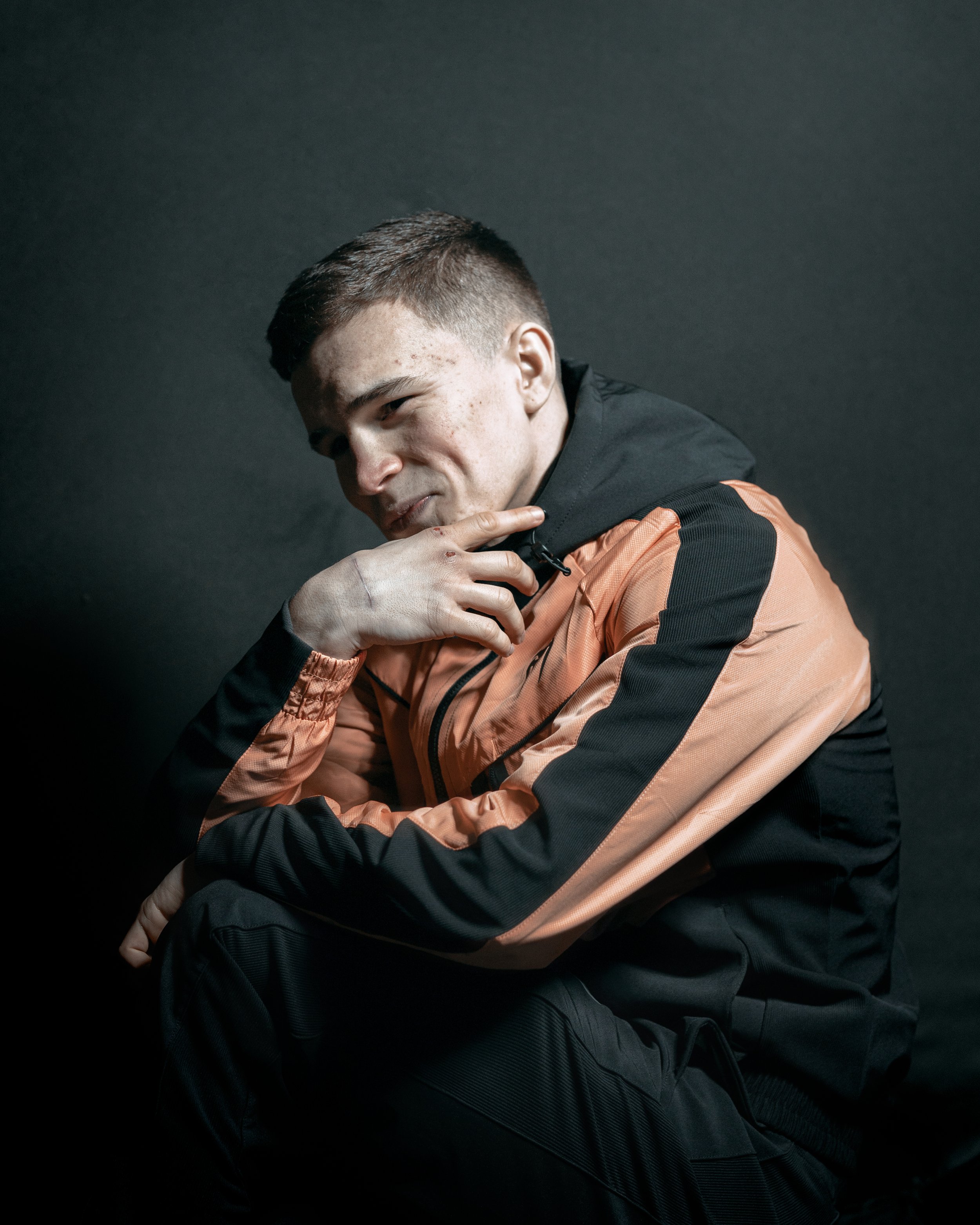 Young man in orange and black jacket sitting against a dark background, looking thoughtful.