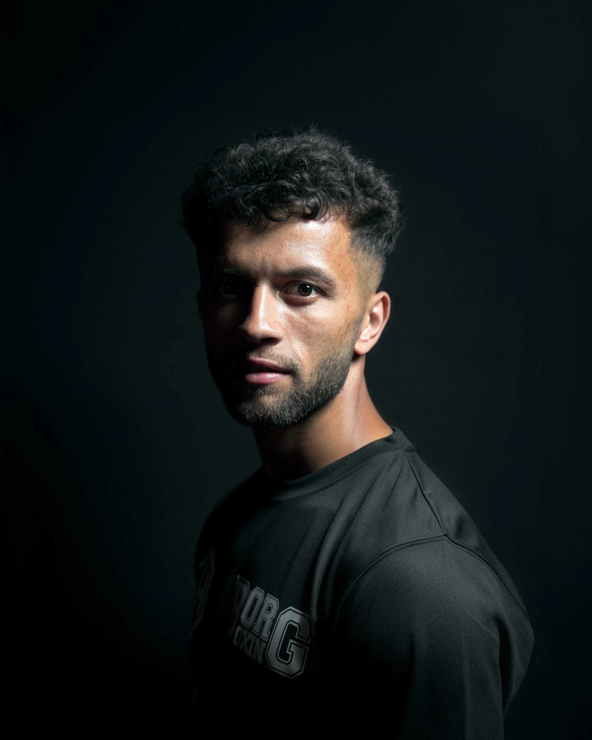 A young man with curly hair and a beard wearing a dark shirt against a black background.