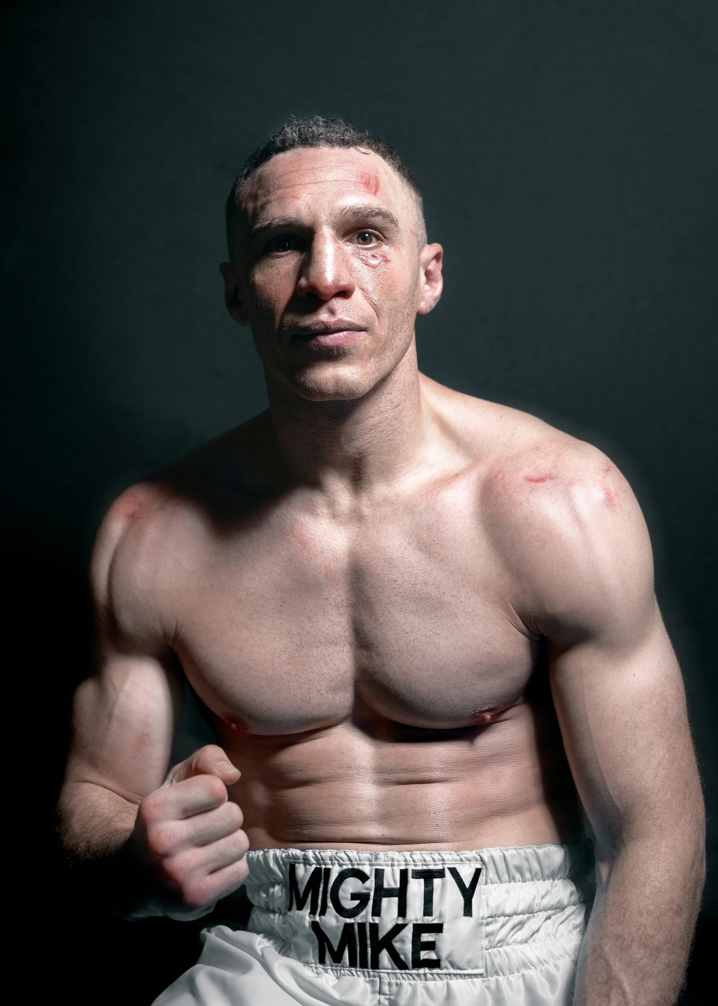 A shirtless male boxer with a determined expression, standing against a dark background, wearing white boxing shorts labeled 'MIGHTY MIKE', with visible injuries and muscle definition.