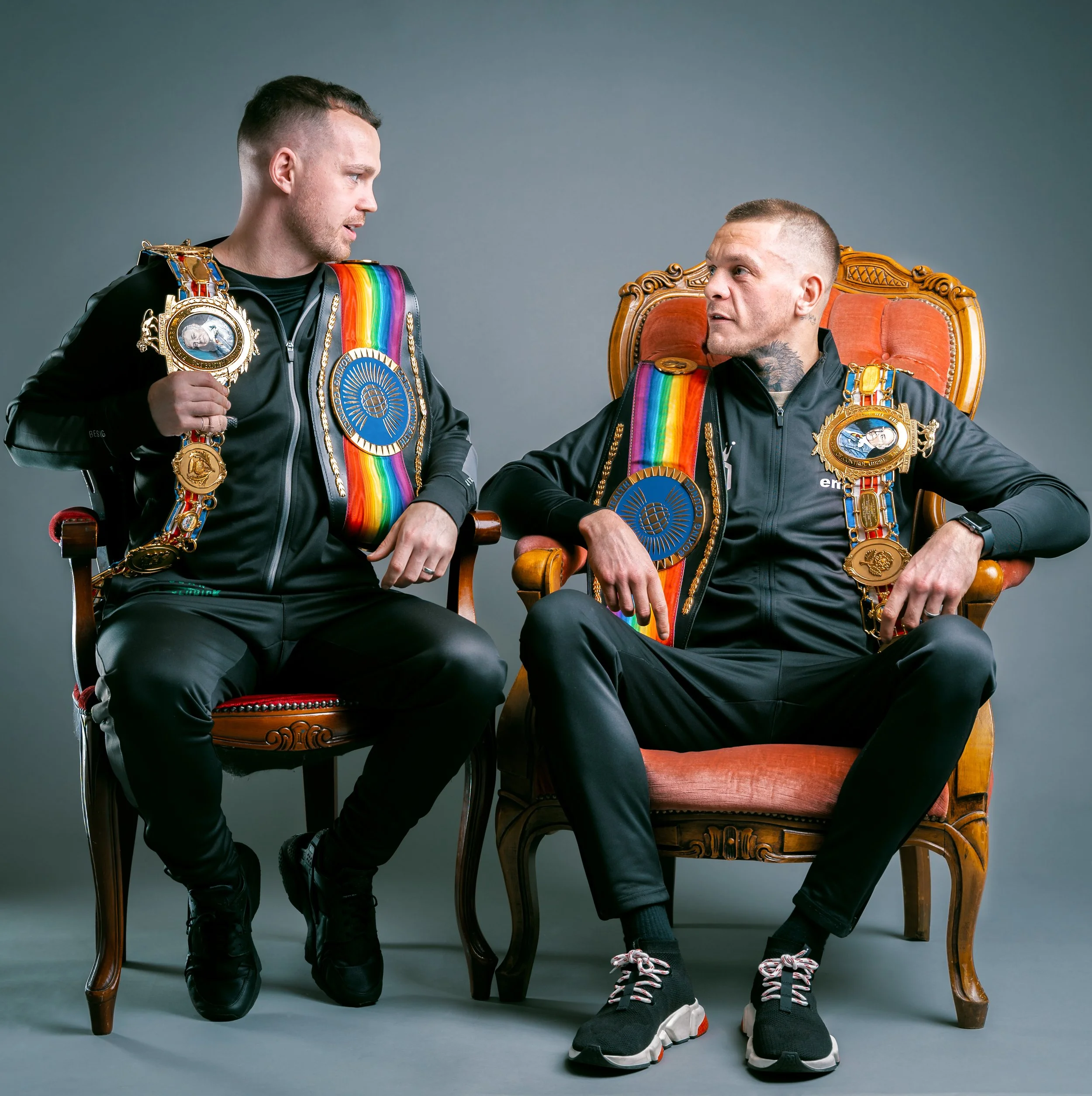 Two men in black athletic clothing sit and stand on antique chairs, wearing rainbow-colored sashes and large medals around their necks. They are engaged in conversation against a plain gray background.