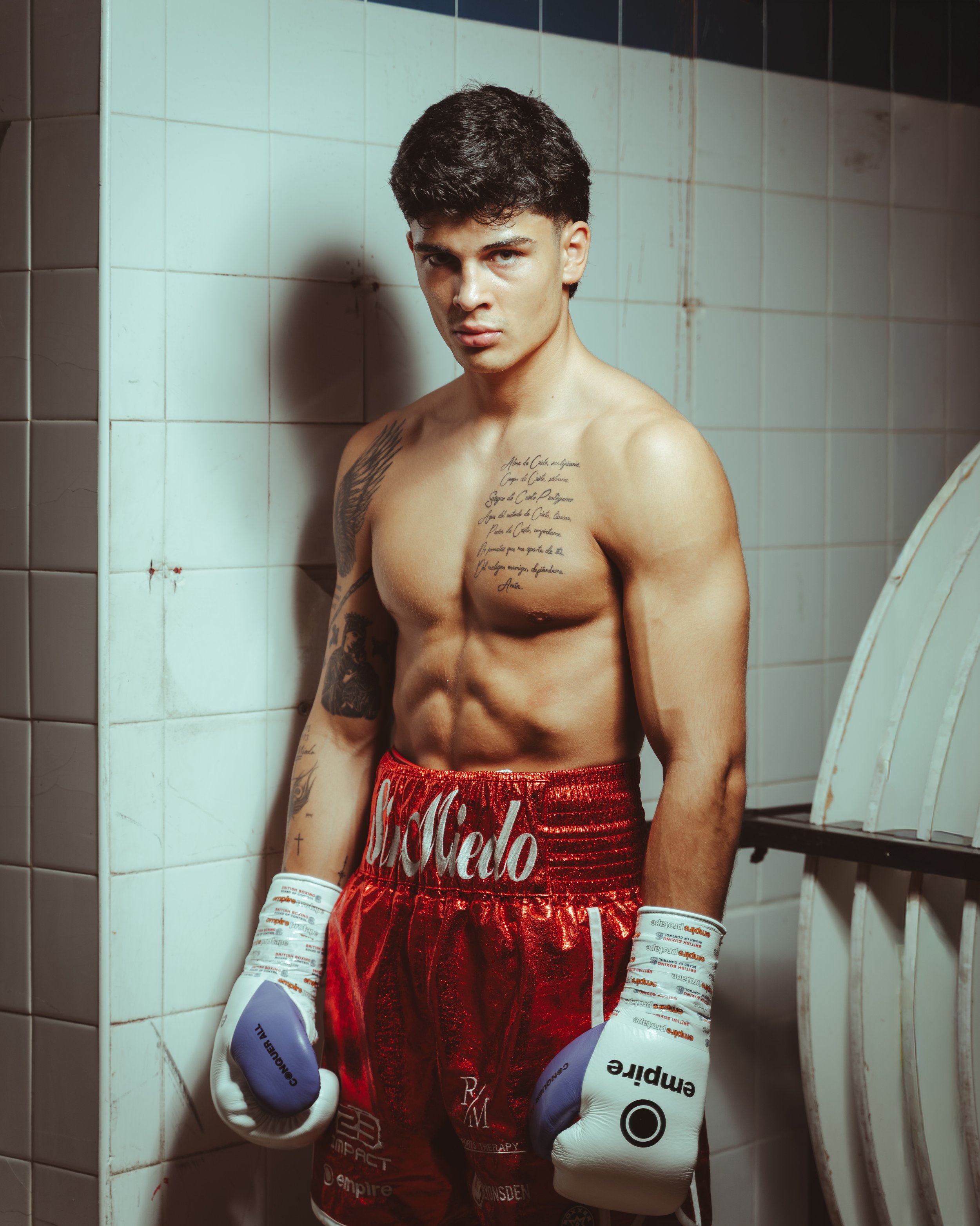 A shirtless male boxer with tattoos, wearing red boxing shorts and white boxing gloves, standing in a tiled room with a serious expression.