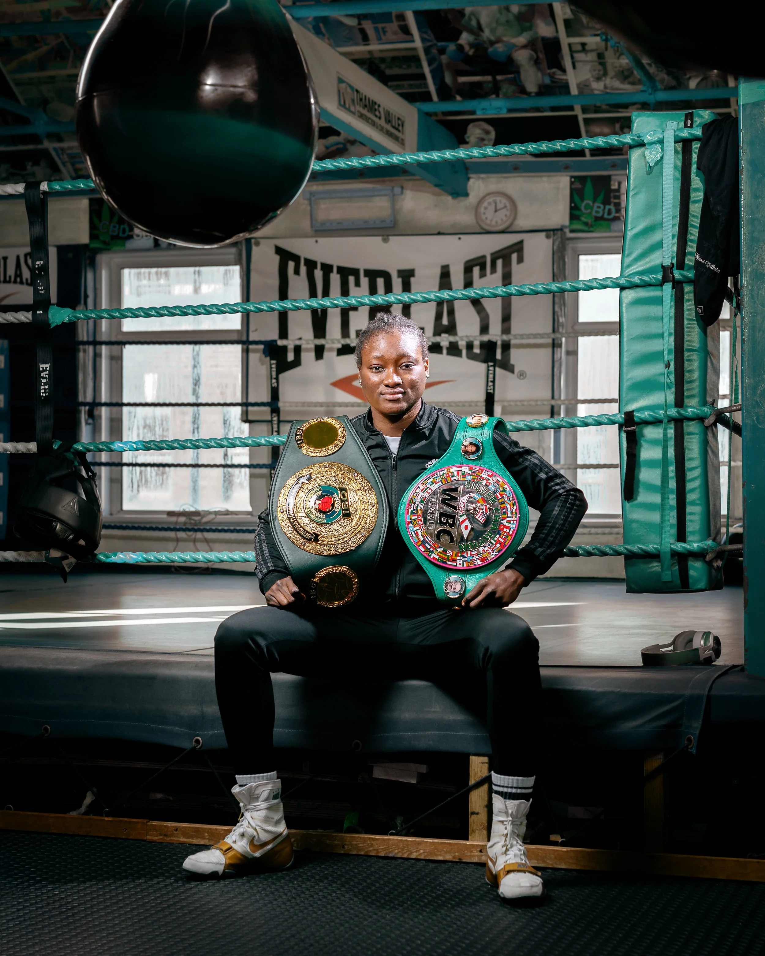 A female boxer sitting on a boxing ring step, holding two championship belts, in a gym with boxing equipment and a large 'EVERLAST' banner in the background.