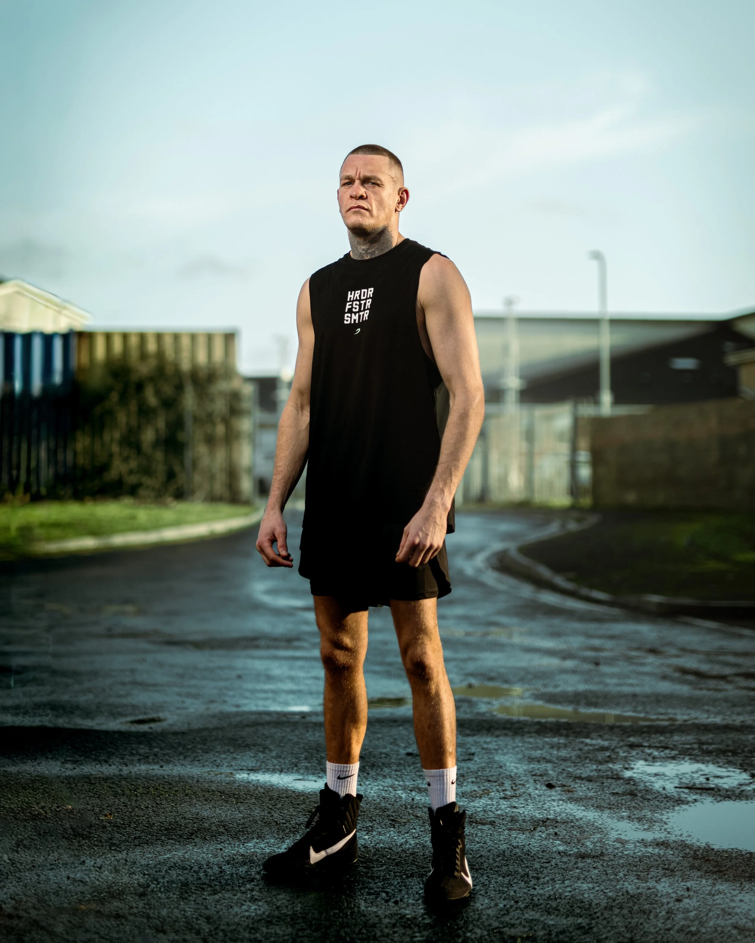 A young man with tattoos wearing a black sleeveless athletic shirt, black shorts, white socks, and black Nike sneakers. He stands on a wet road with an overcast sky, residential buildings, and a fence in the background.