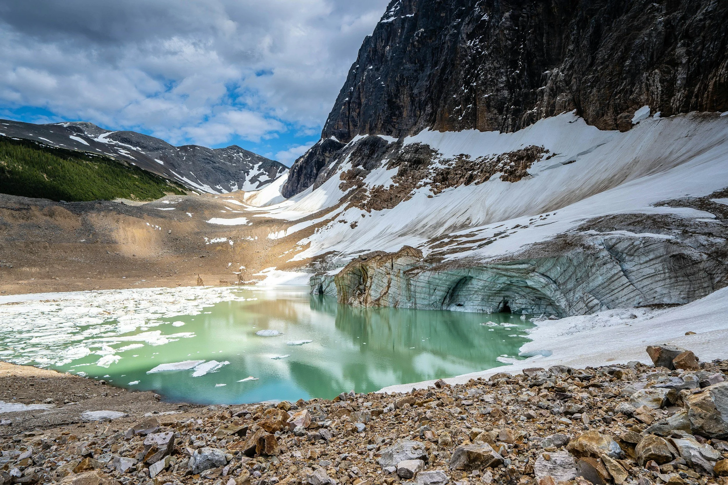 A mountain landscape with snow-covered peaks, a glacier at the Columbia Icefields with tunnels, and a small turquoise glacial pond surrounded by rocks.