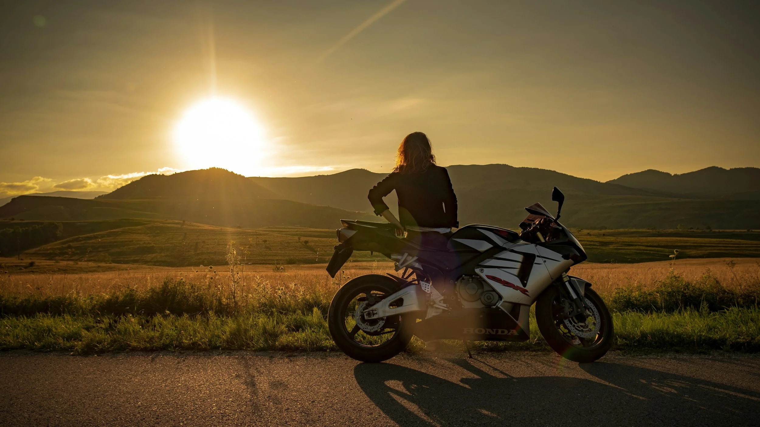 A woman stands next to a silver and black Honda CBR sportbike on an empty road during a sunrise, looking out over a scenic valley and rolling hills.