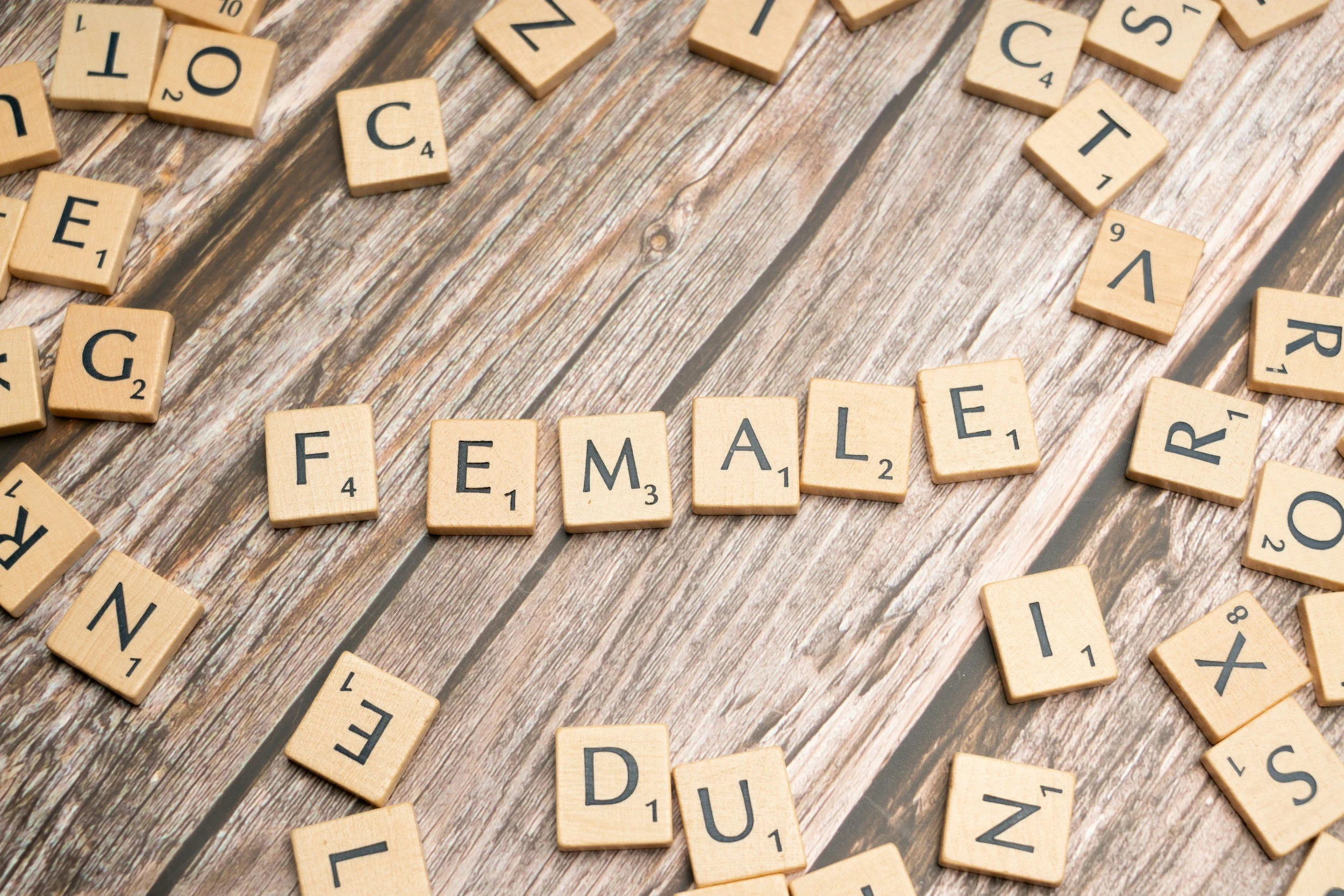Wooden letter tiles spelling out the word FEMALE arranged horizontally across a rustic wooden table surrounded by scattered random letters.