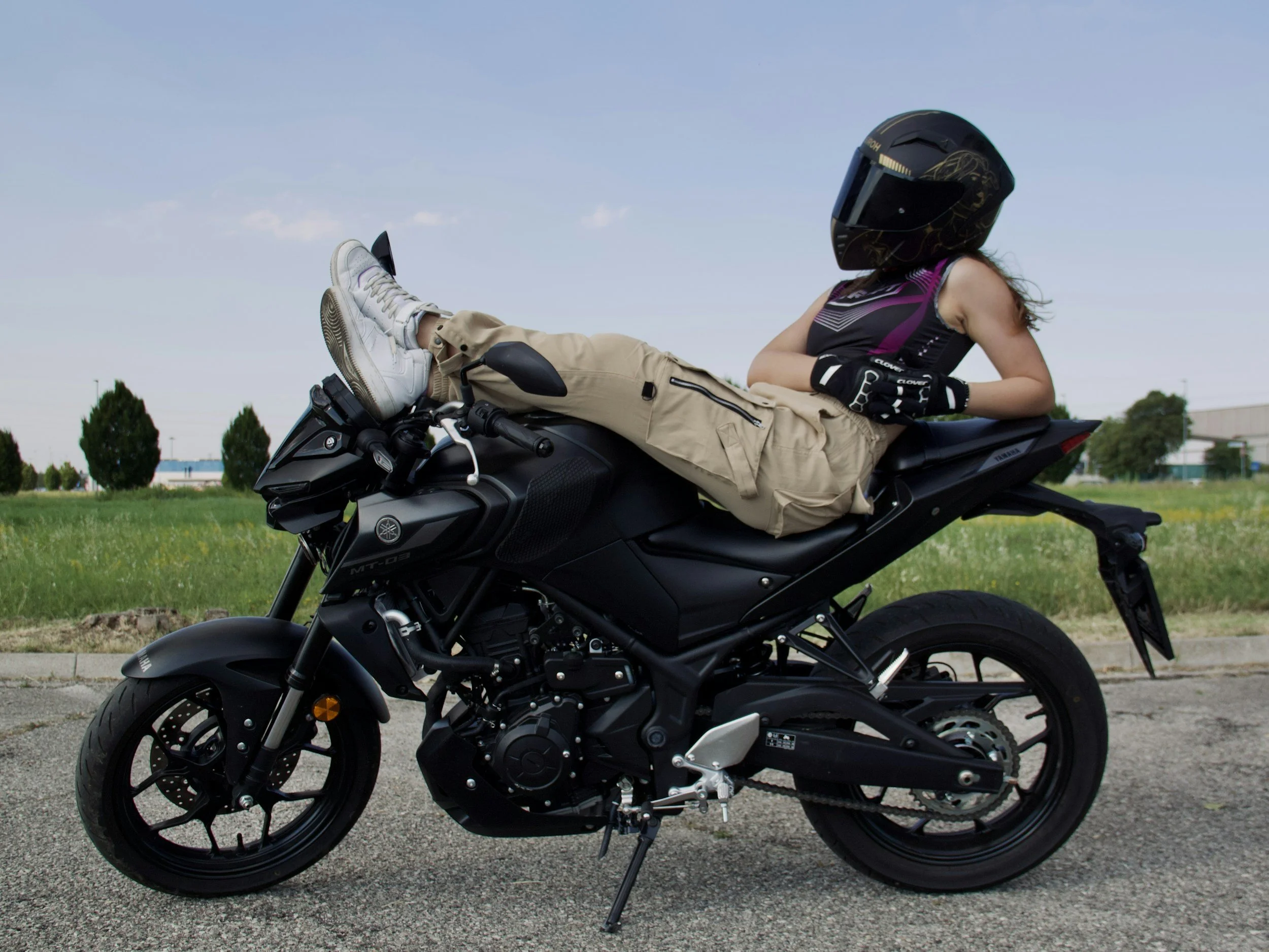 A woman wearing a helmet and cargo pants relaxes by leaning back and resting her feet on the handlebars of a black Yamaha MT-03 naked motorcycle.